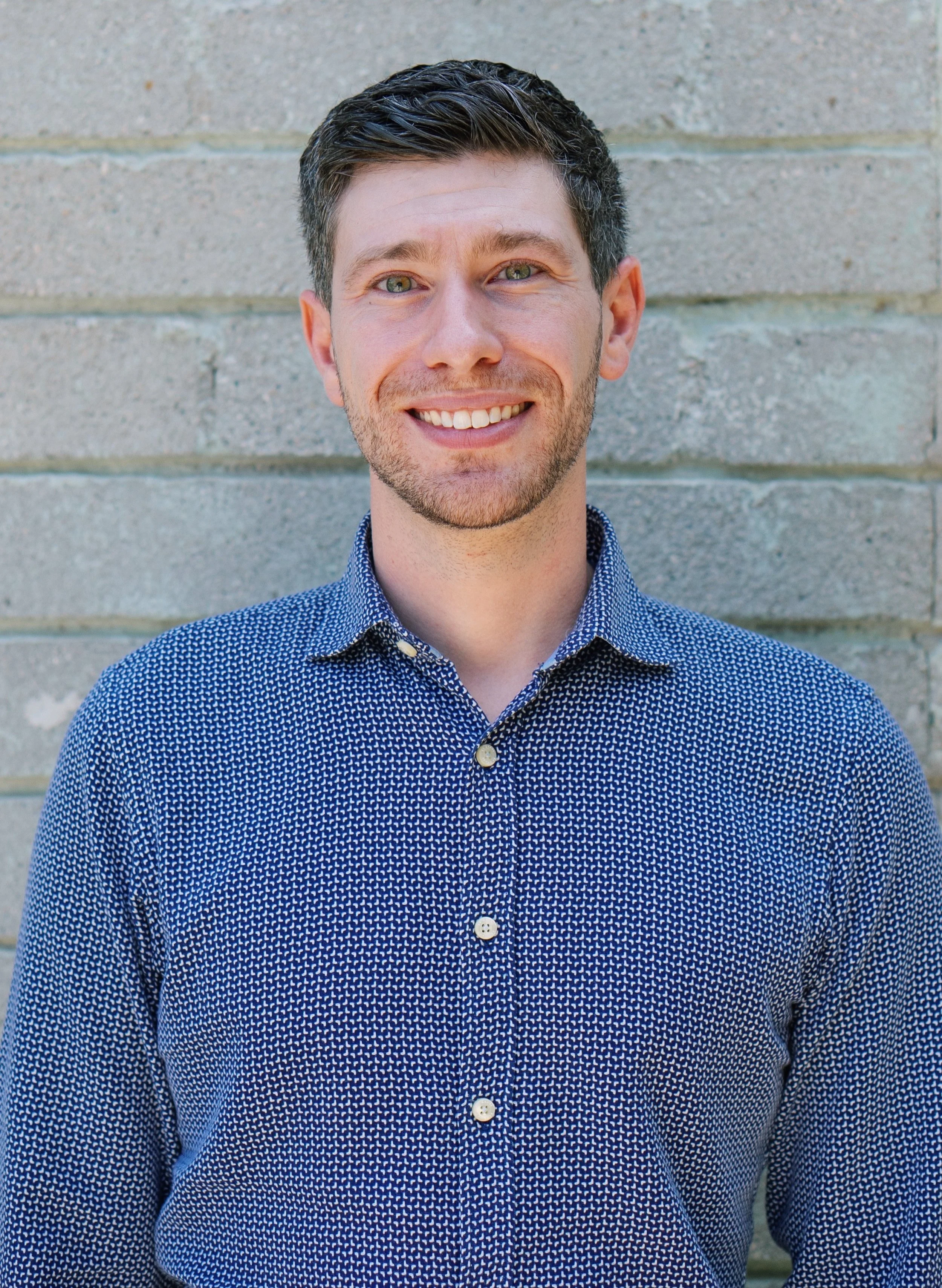 A white man with light brown hair, green eyes, thin beard, wearing a blue-patterned button up and smiling.