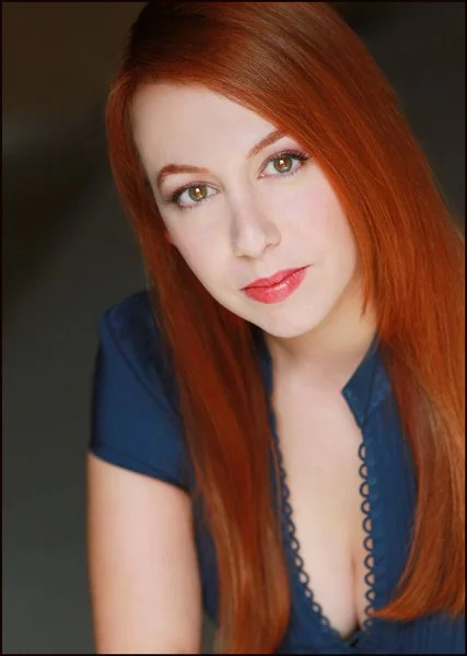 A headshot of a white cuban woman with red hair, wearing a navy dress.