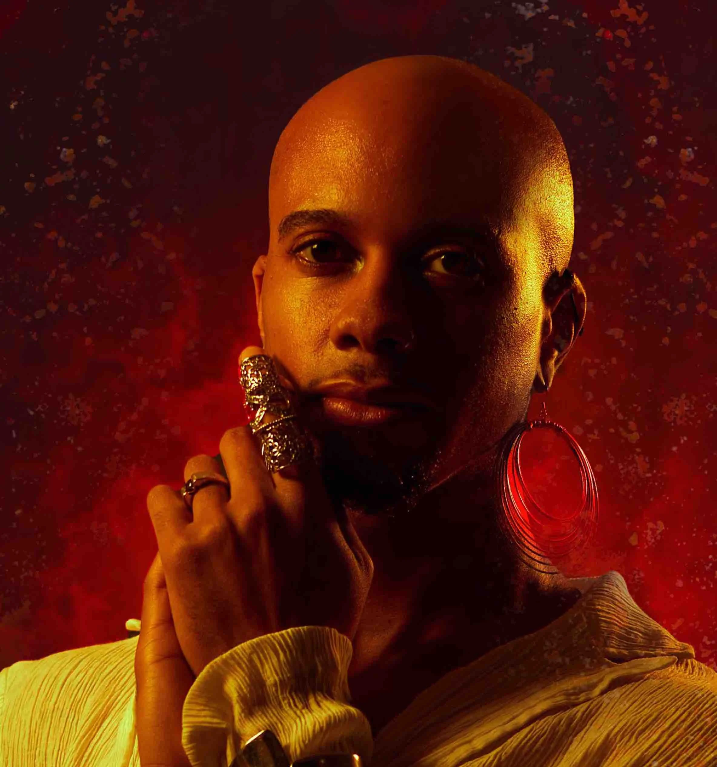 A bald, non-binary Black person looks into the camera with a slight smile. They have a mustache, and are wearing a long-sleeve, flowy, pale yellow shirt and large hoop earrings.