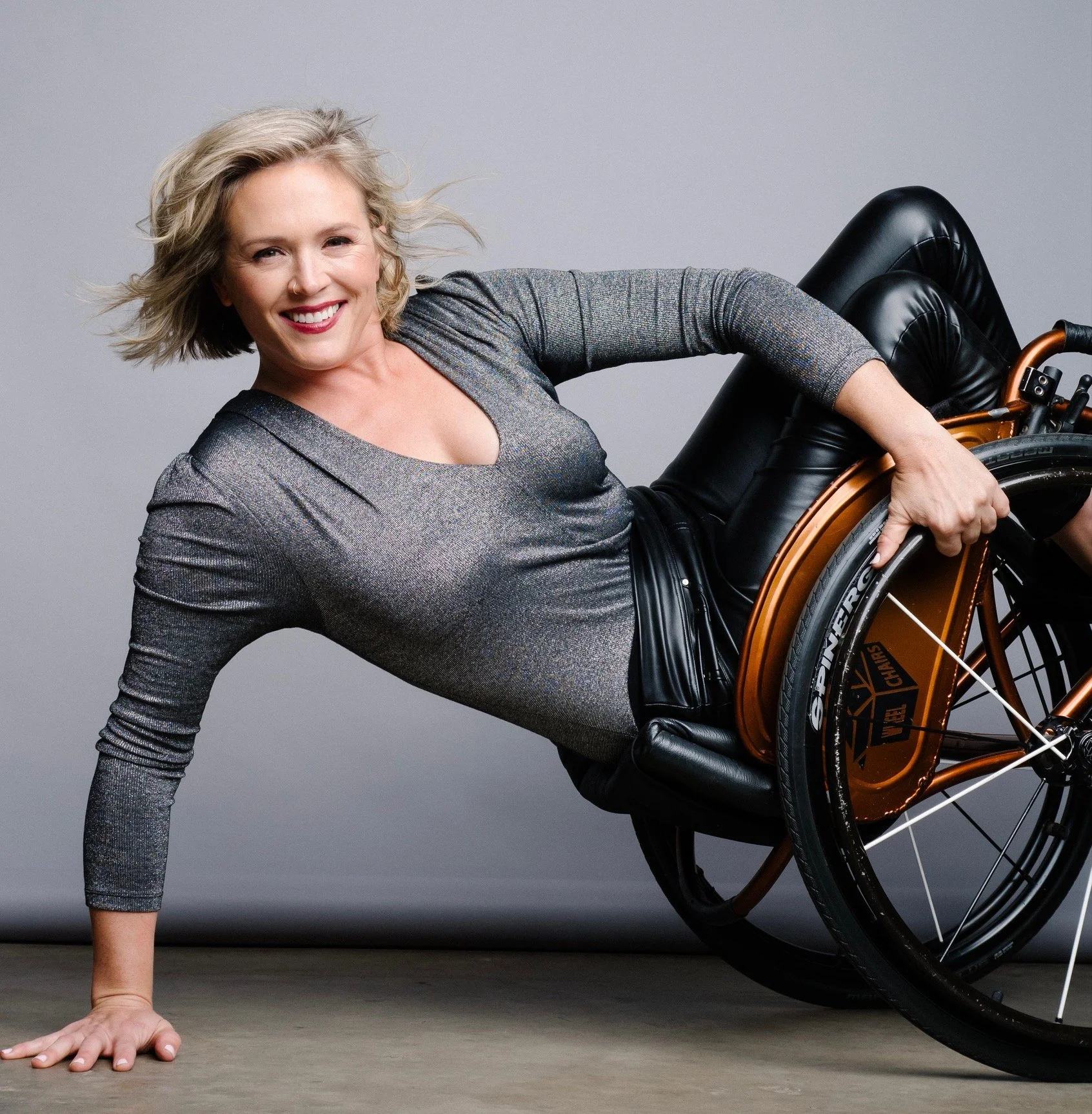 Jamey Perry, a blonde white woman in a sparkly top and leather pants poses in a "layback" position in a bronze wheelchair, with her right hand on the ground and the chair's casters in the air.