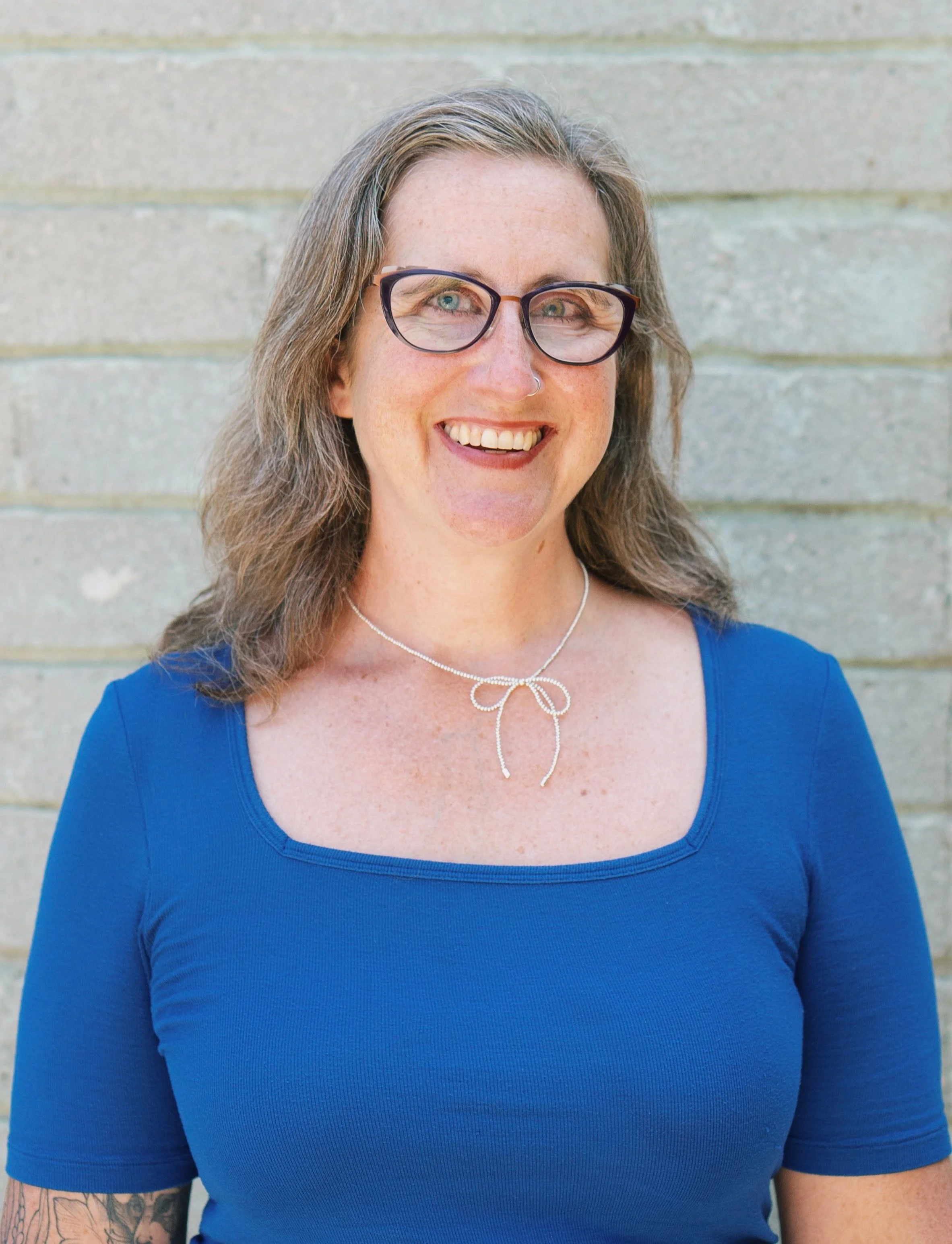 A smiling white woman with long brown hair, a nose ring, and thick glasses in a white t-shirt, against a gray background.