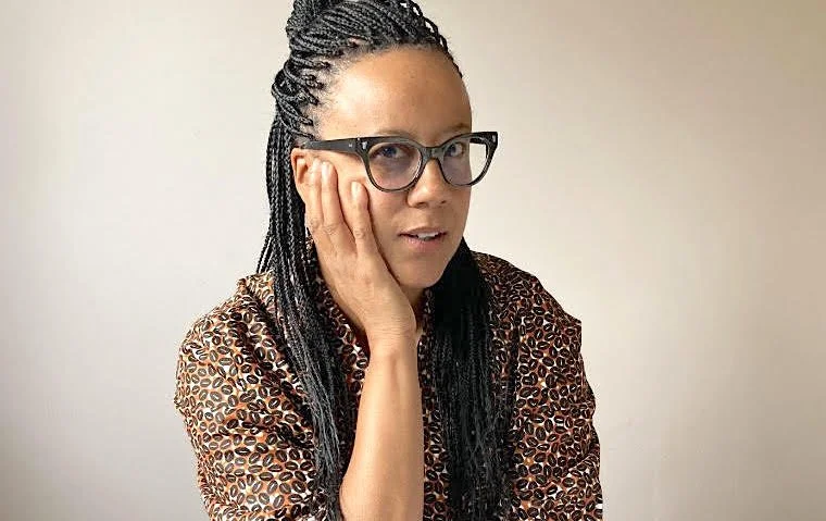 A bi-racial woman wearing her hair in long braids. She is wearing black framed glasses and a brown jacket with a coffee bean print. Her face is resting in one hand.