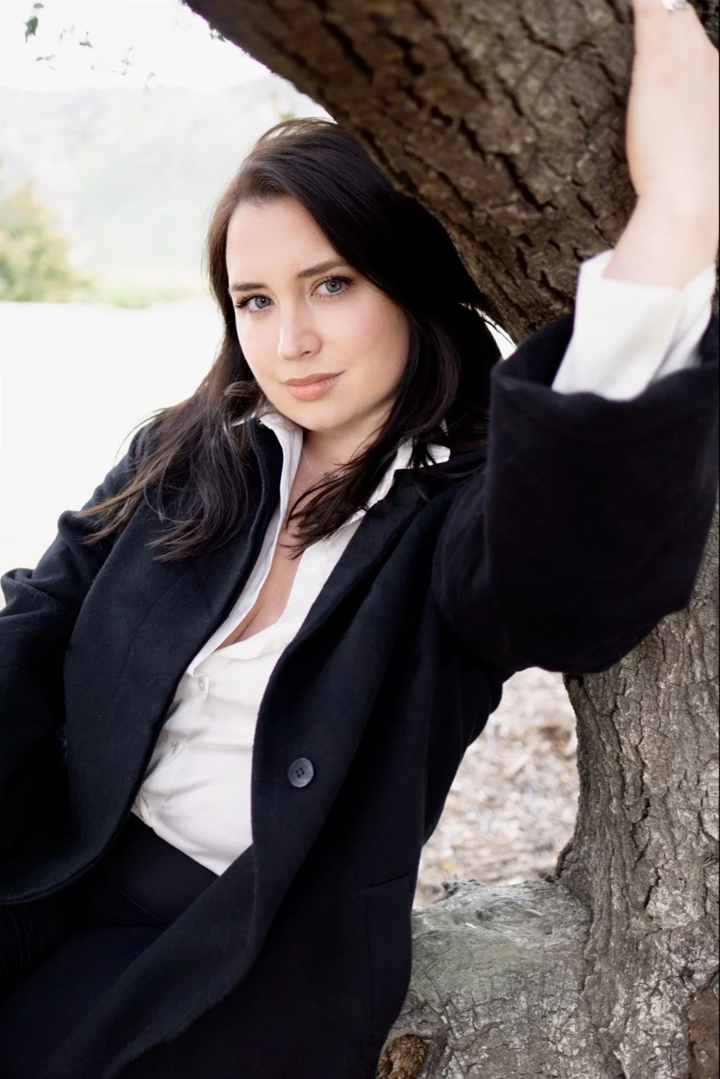 A white woman with long brown hair and blue eyes, wearing a white shirt and black coat, and smiling.
