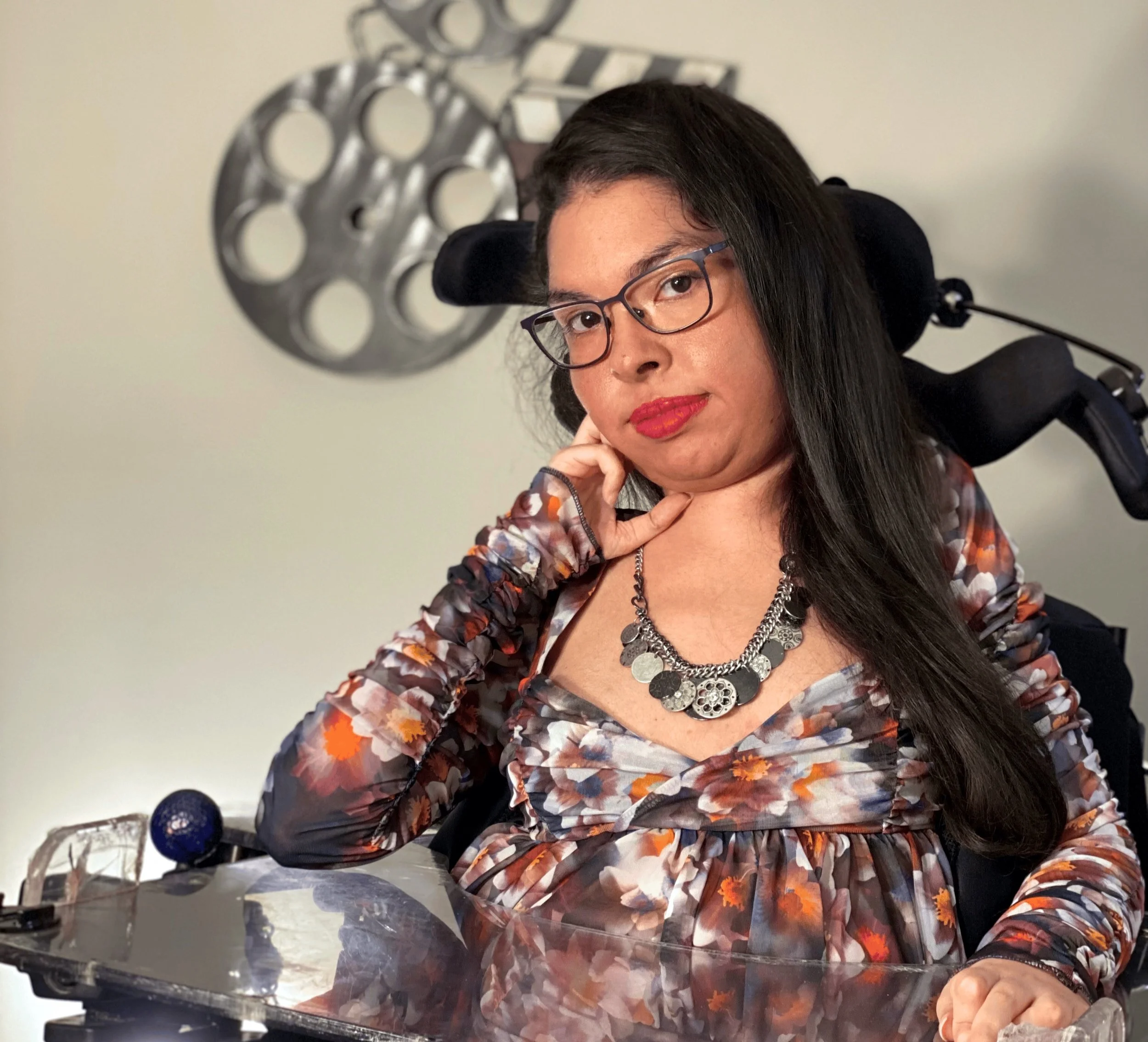 A brown gal with side swept long black hair and a hot pink smirk rests her cheek against her hand and wheelchair headrest. She’s parked in front of a film reel art piece on the wall behind her.