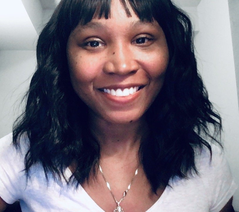 A black woman with black hair and brown eyes. She is wearing a white t-shirt and is looking at the camera with a smile.