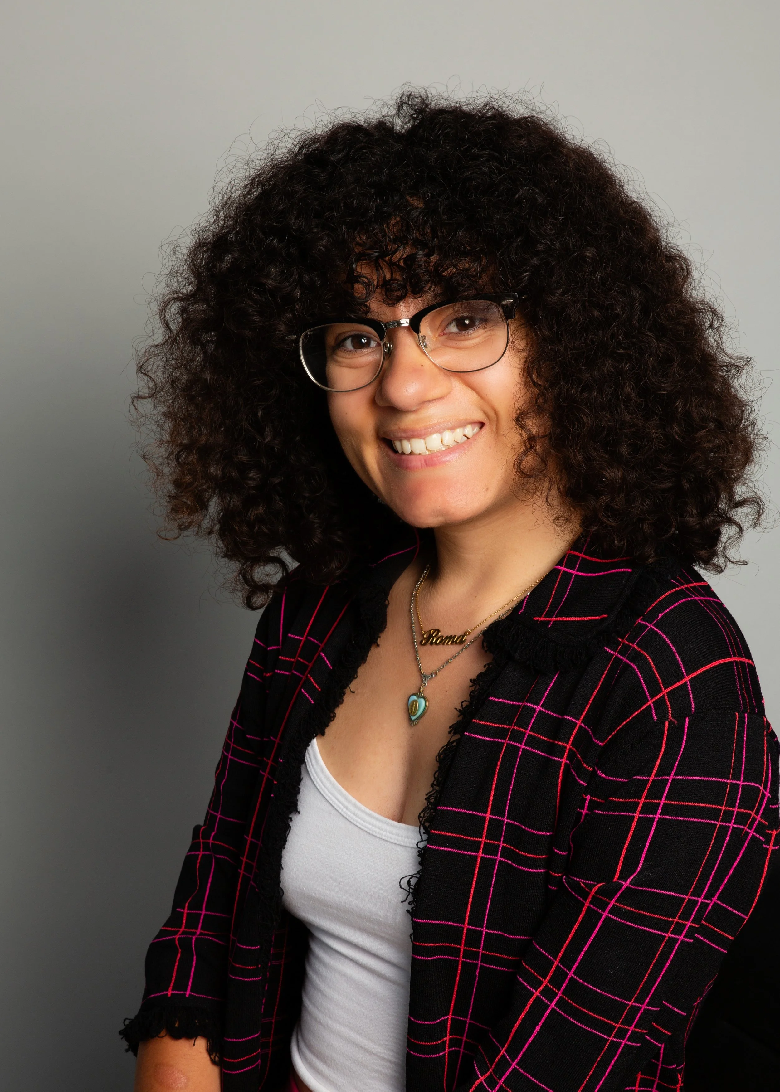 A lightskinned black woman with shoulder-length curly brown hair, smiles at the camera against a grey backdrop. She is wearing a white shirt, several necklaces, and a pink and black plaid cardigan.