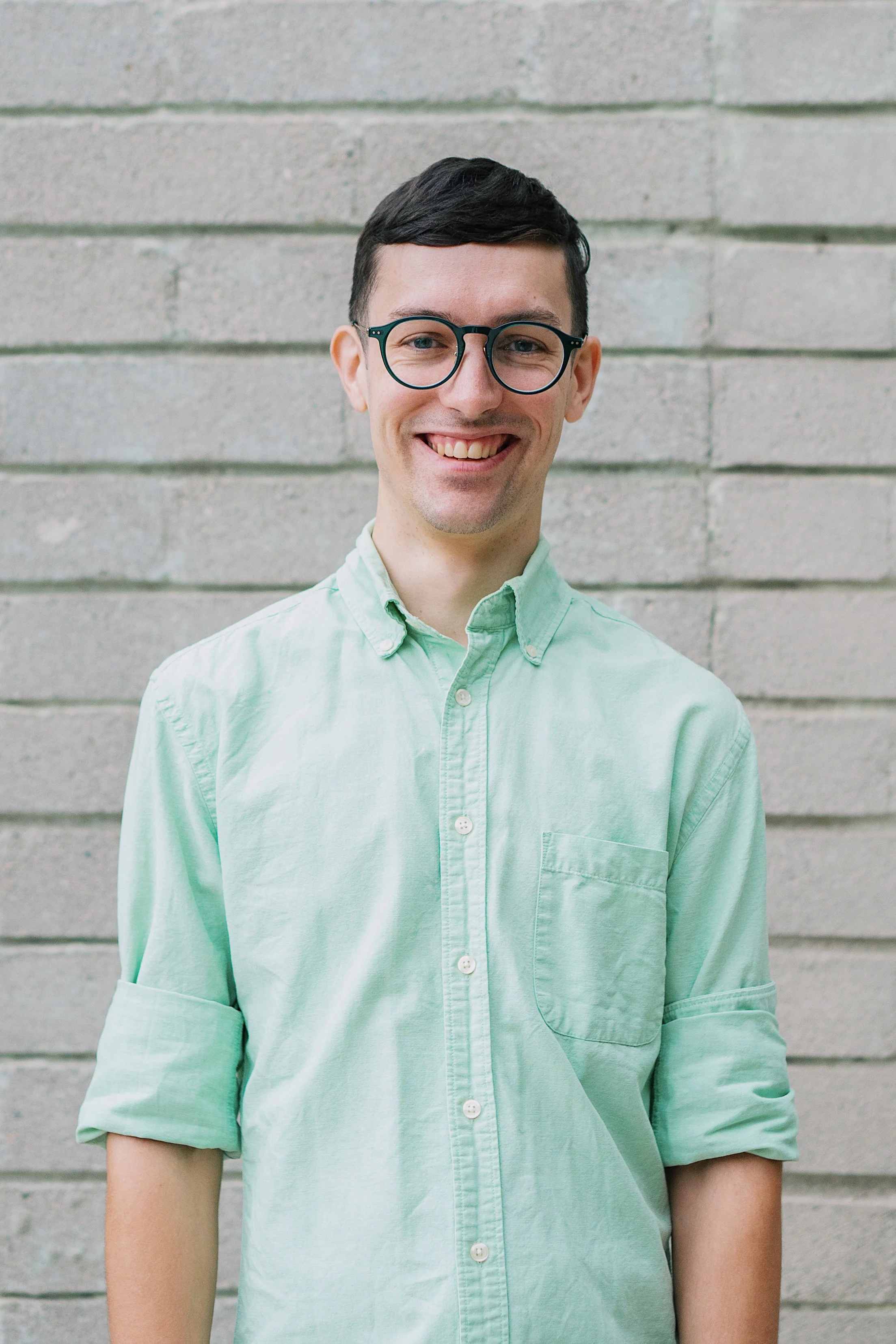 A headshot of a man with dark hair, wearing blue glasses and a light blue button-up shirt against a grey brick background, looking into the camera