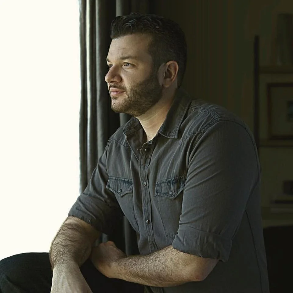 An Iranian-American man with short brown hair, brown eyes, a brown beard, and wearing a gray, long-sleeved shirt.