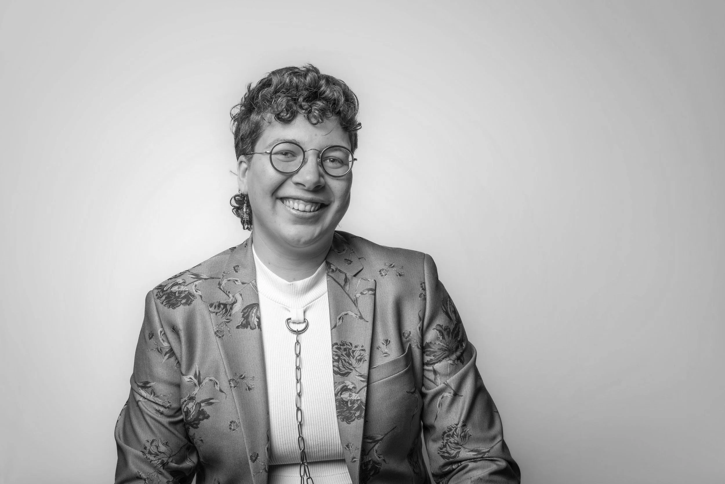 A black and white photo of a person with curly hair and glasses smiling and looking directly to camera.