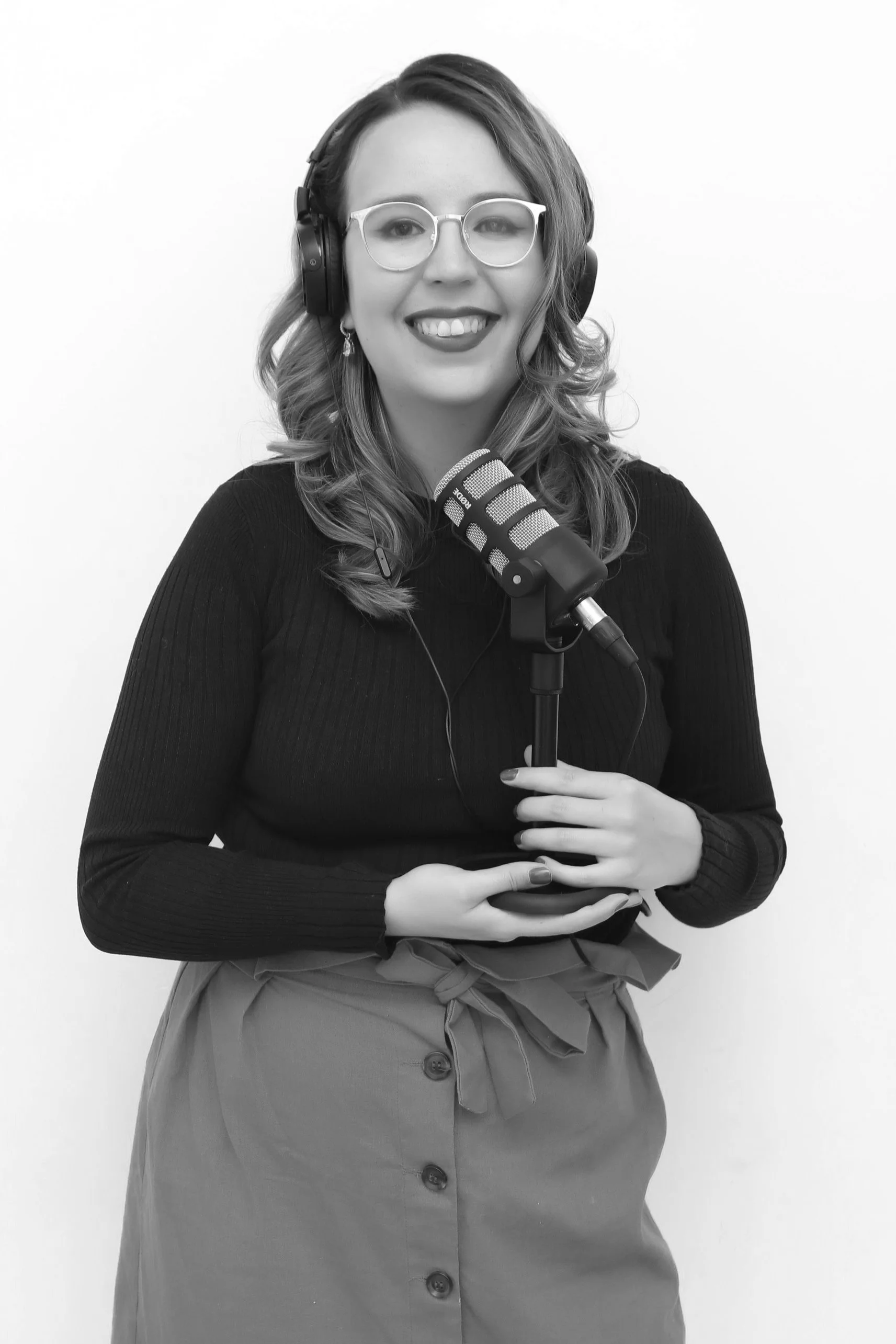 A black and white photo of a person with a black shirt, glasses, and grey skirt holding a microphone and wearing headphones while smiling directly to camera.