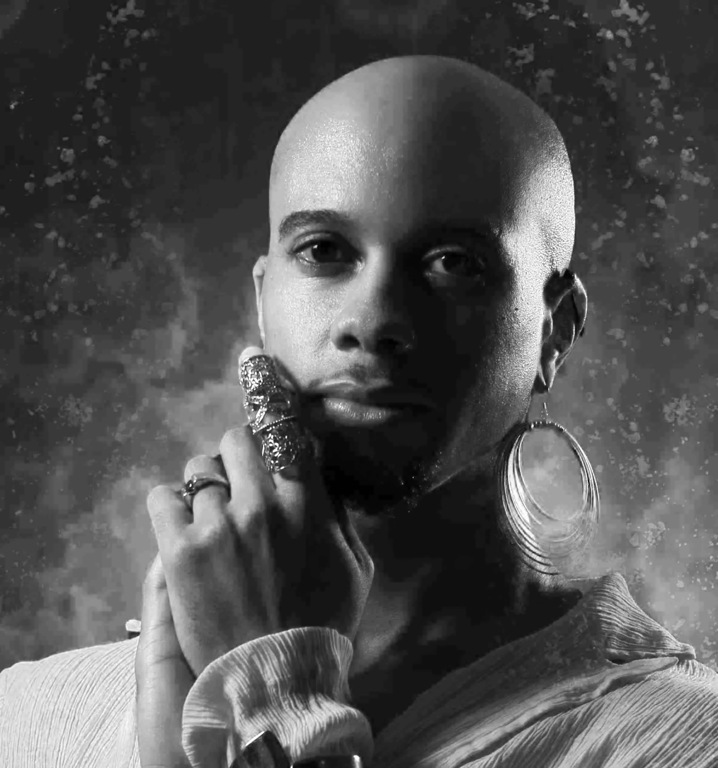A black and white image of a person with jewelry on their hand and ear, looking towards the camera with their hand placed on their chin.