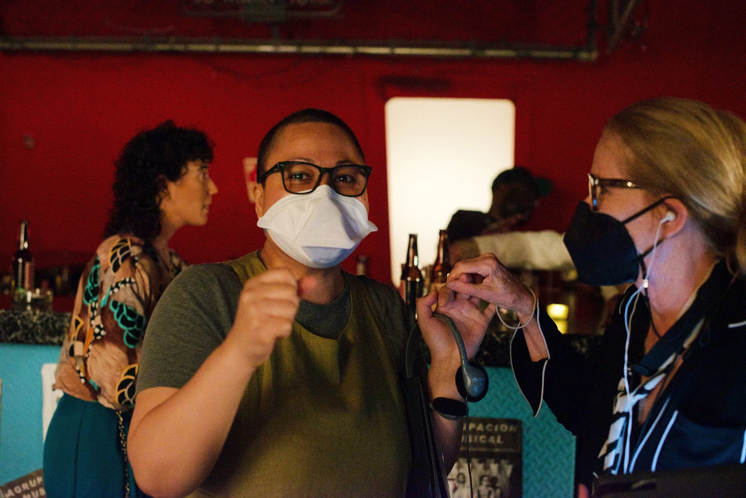 A director with glasses and a mask on looking directly into camera while a crew member with a mask on looks left towards them. There are people in the background and a red wall.
