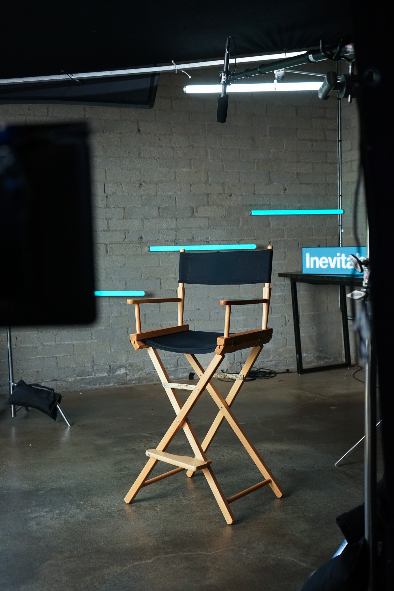 A photo of a director's chair, sitting in the middle of a set with grey brick walls seen in the background. There is camera equipment surrounding the chair and a table behind it with an Inevitable Foundation sign partially seen.