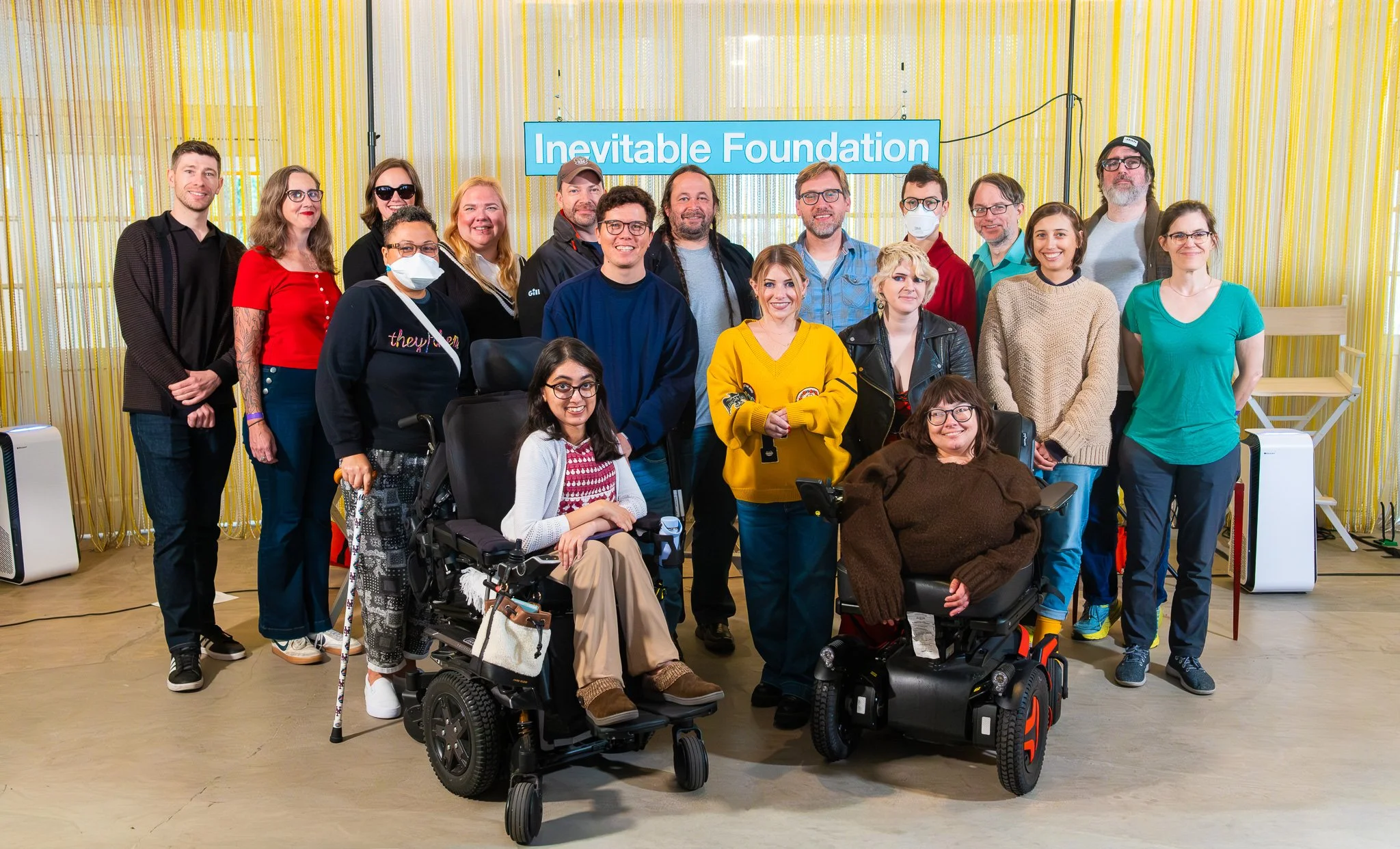 A group of Inevitable Foundation's Accelerate fellows, standing in front of an Inevitable sign in a brightly lit room. Fellows are seen smiling at the camera, some with masks and mobility aids and some without.