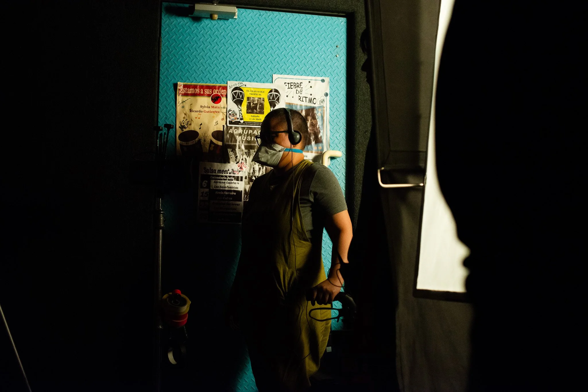 An image of a person directing a film, wearing headphones and a mask in front of a teal background with posters on it.