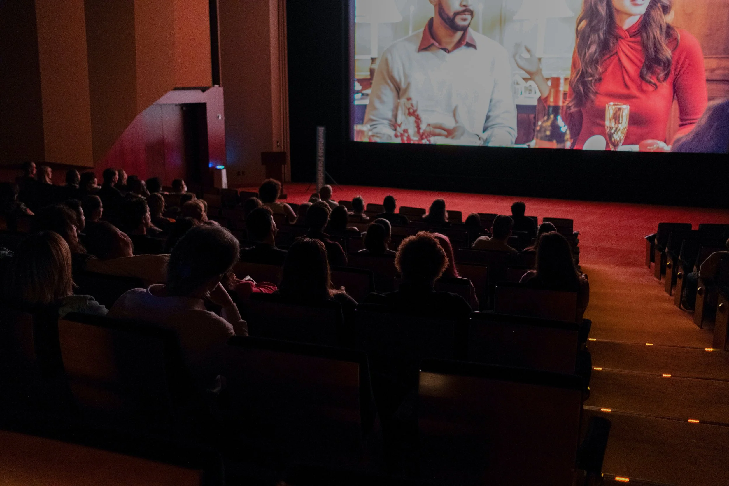 A photo of a movie theater with the silhouette of an audience, as they watch a film on screen.