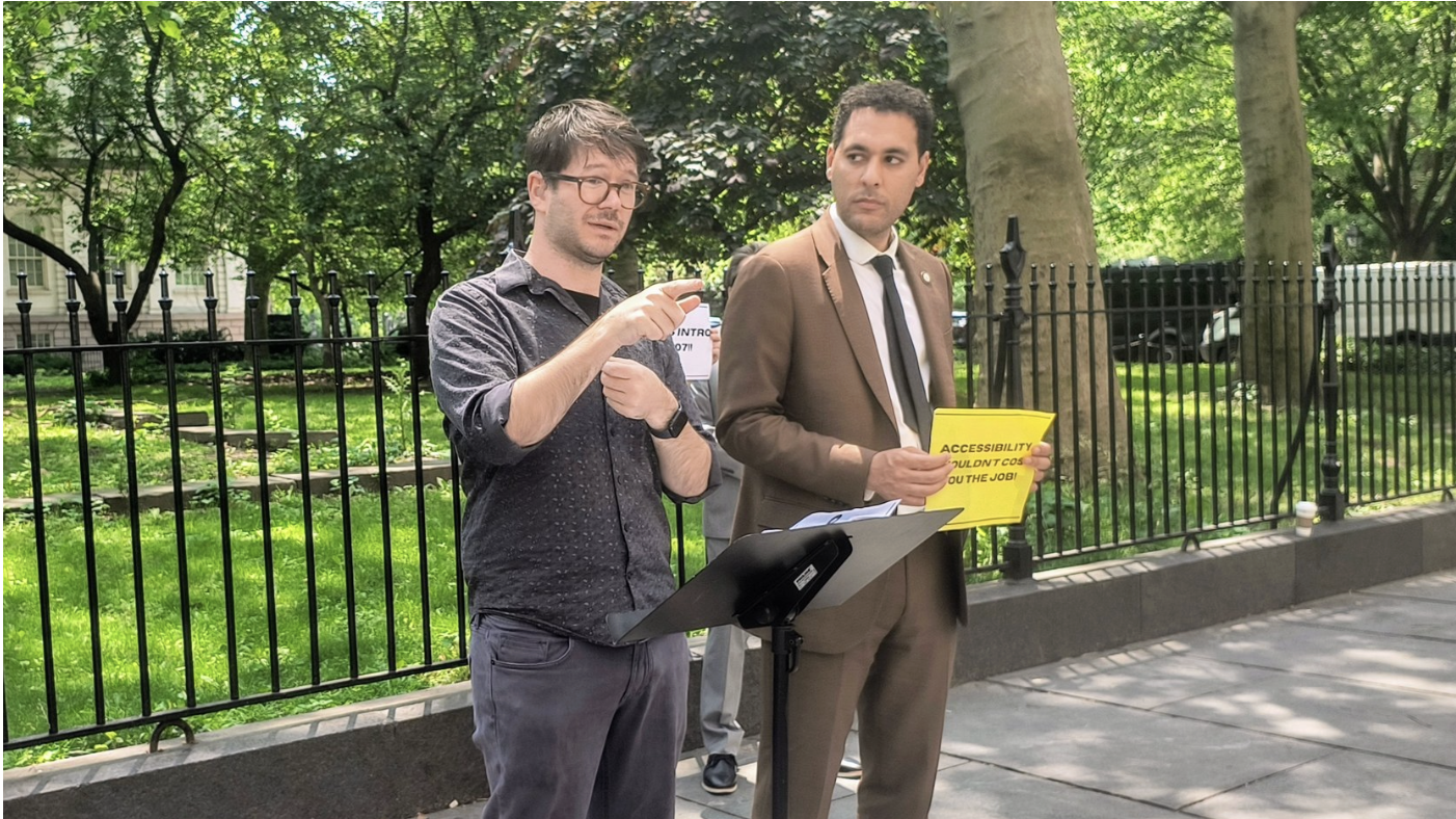 A photo of two people in front of a fence speaking. One is wearing a collared shirt and glasses, and the other is wearing a brown suit and holding a yellow piece of paper. There is a music stand in front of them.