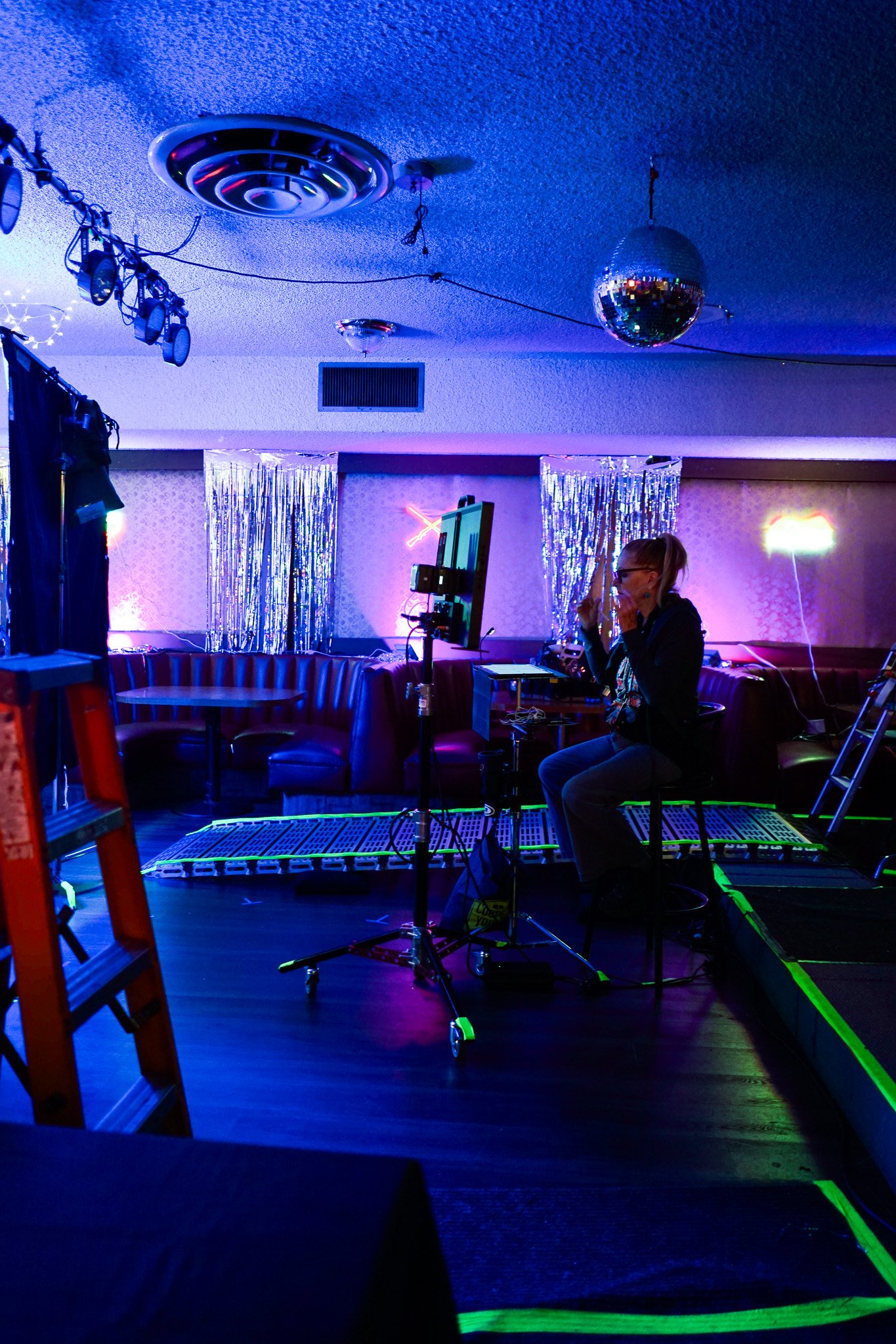 A photo of a camera operator watching a scene being filmed on a set, with silver drapes on the wall in the background and film equipment surrounding the camera and operator.