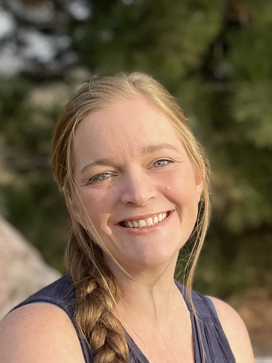 A woman with blonde hair and blue eyes smiling outdoors with green foliage in the background.