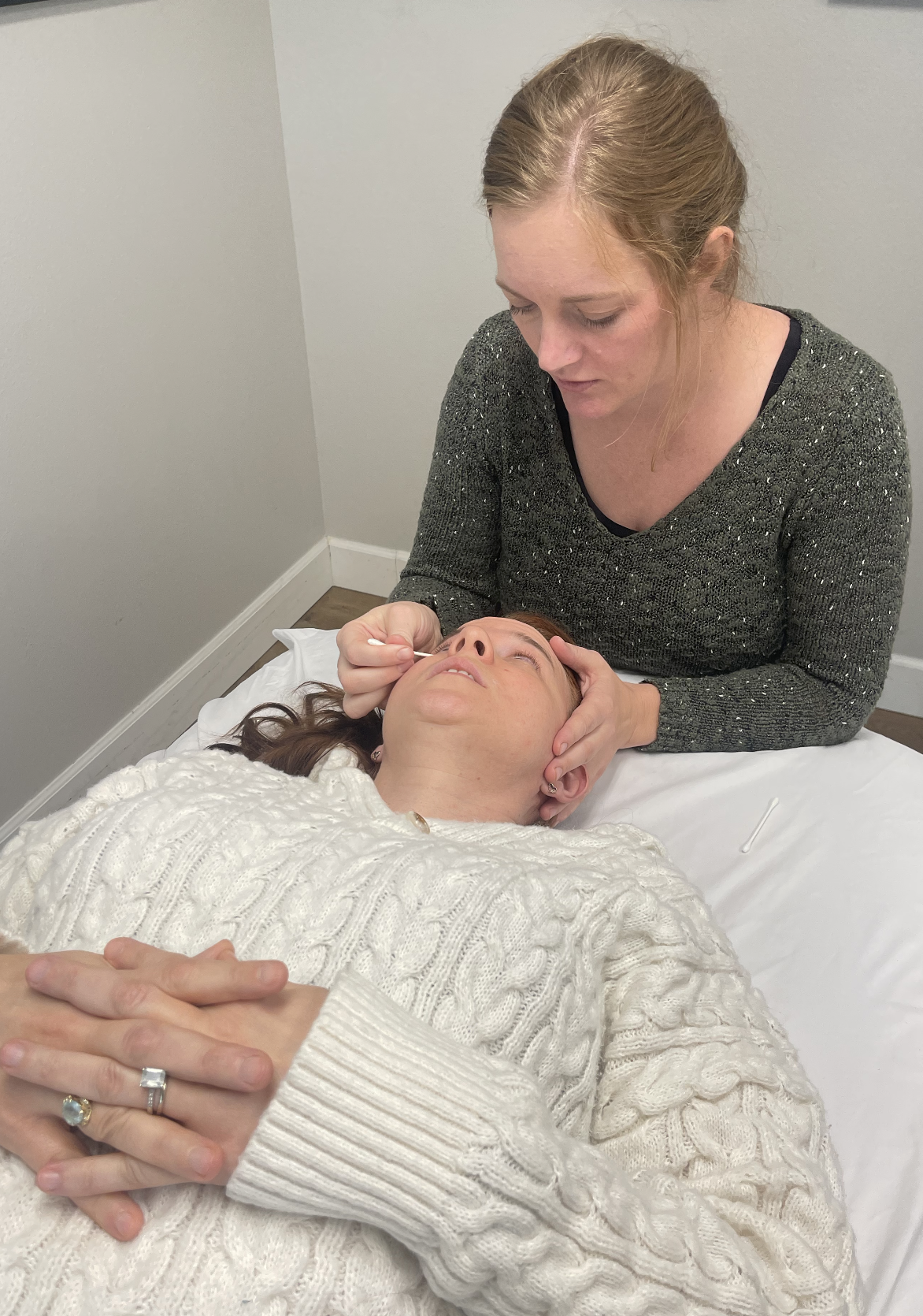 A woman lying on a bed receiving a facial treatment from another woman who is gently holding her face.