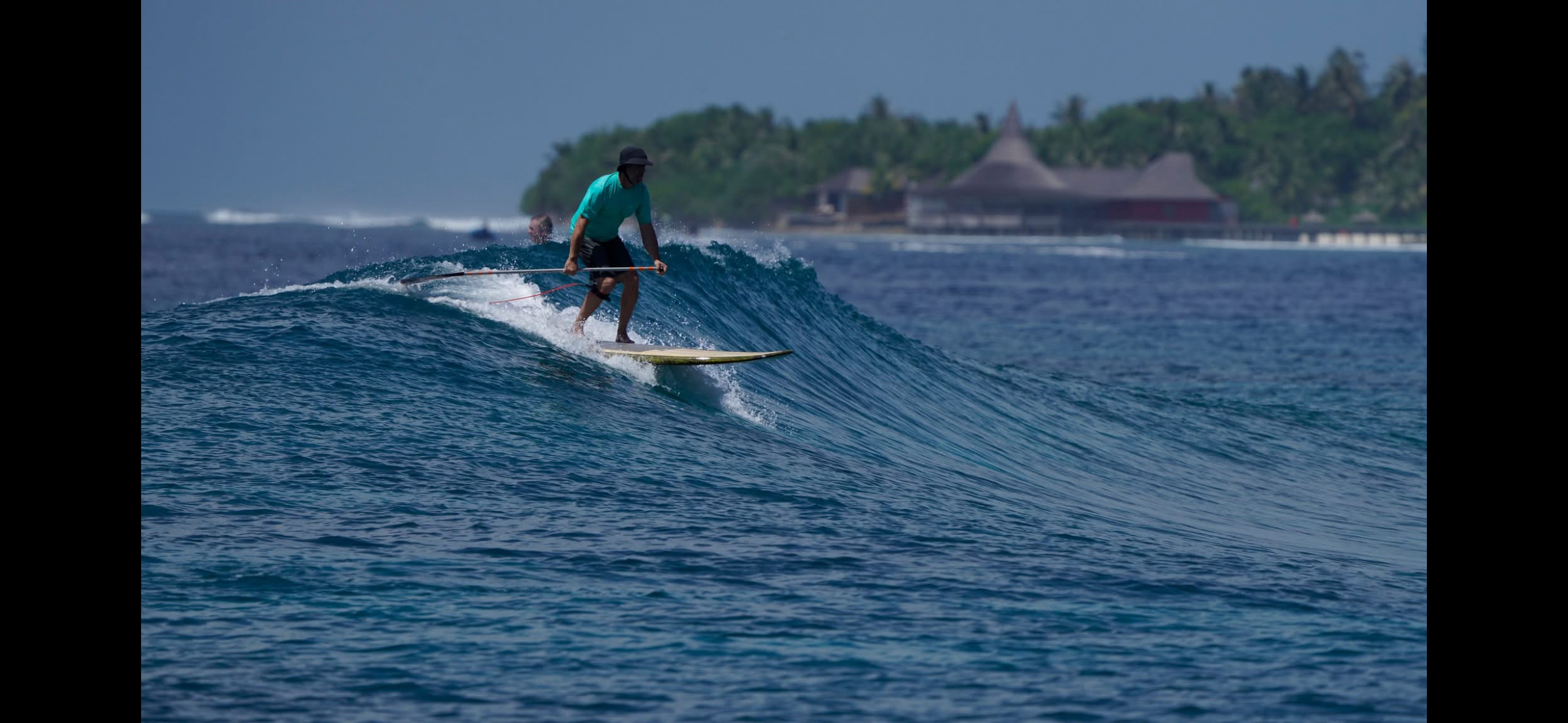 A person wearing a turquoise shirt, black shorts, and a black hat surfing on a blue wave near a tropical shoreline with buildings and lush greenery in the background.