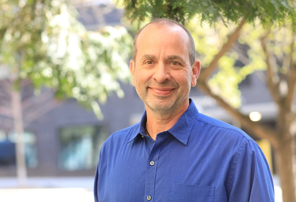 A middle-aged man with a slight smile, wearing a blue dress shirt, standing outdoors with trees and buildings in the background.