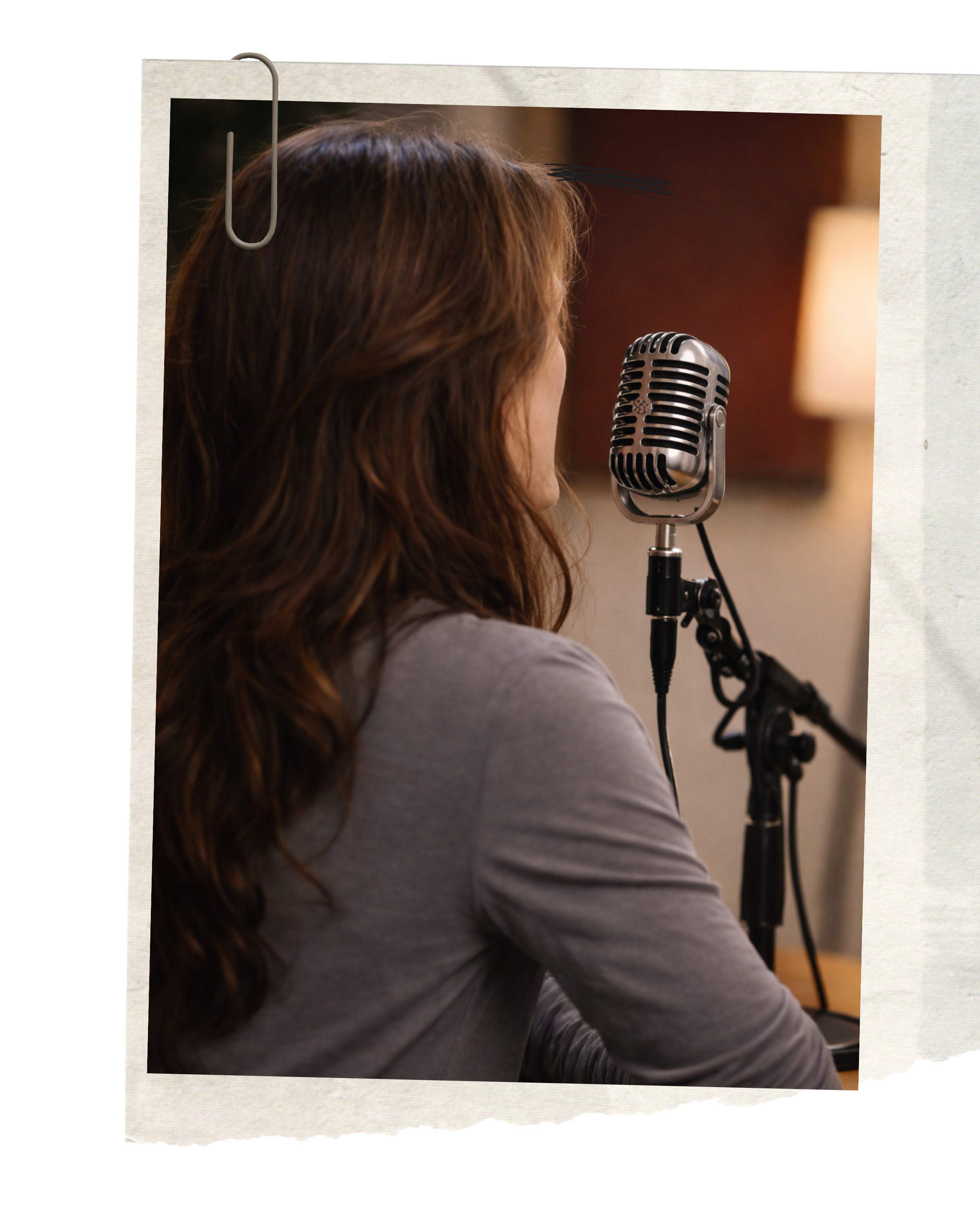 Woman with long, reddish-brown hair speaking or singing into a vintage-style microphone on a stand with a dark background and warm lighting.