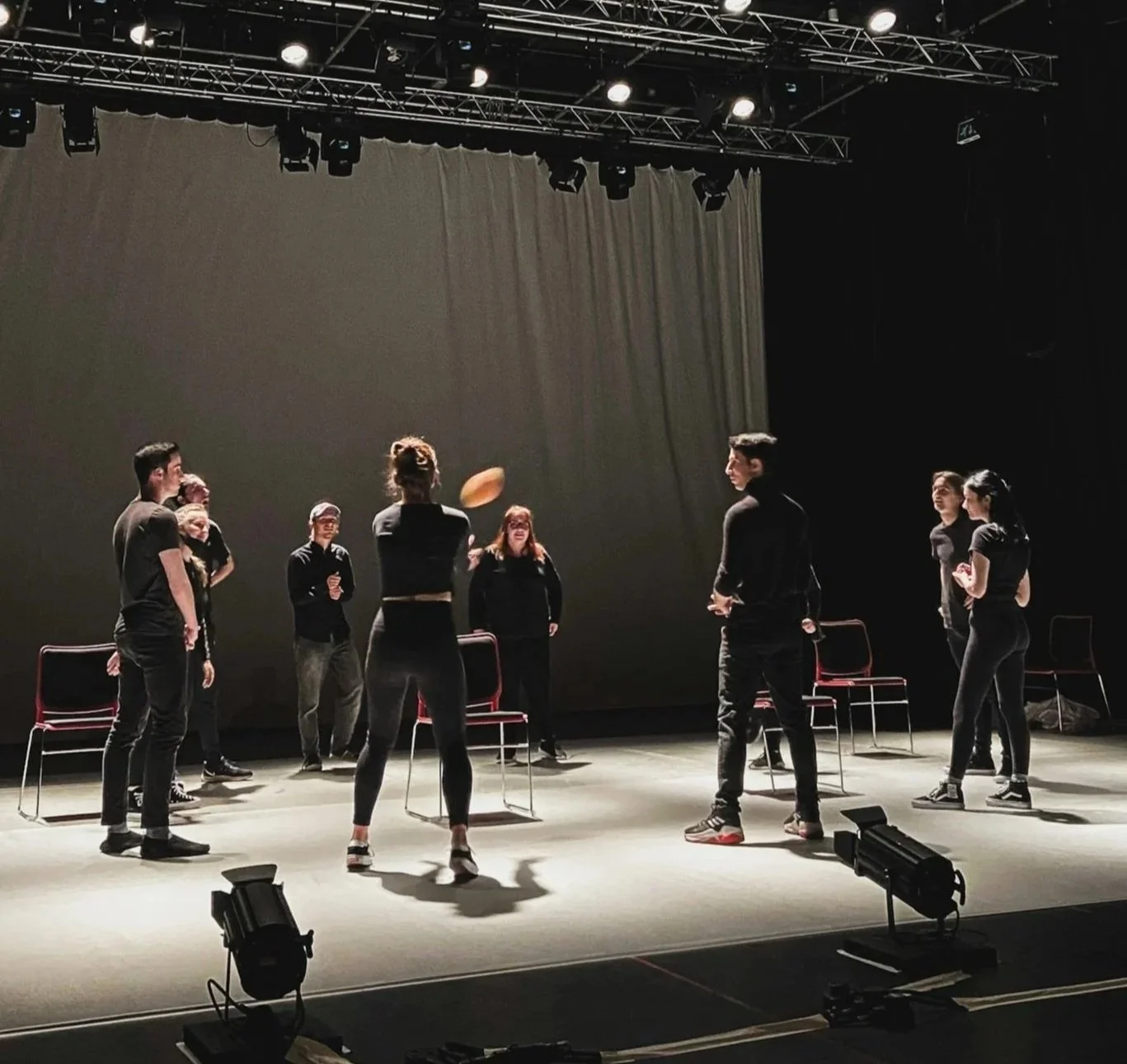 Group of people on stage practicing or performing, with some chairs and stage lighting overhead.