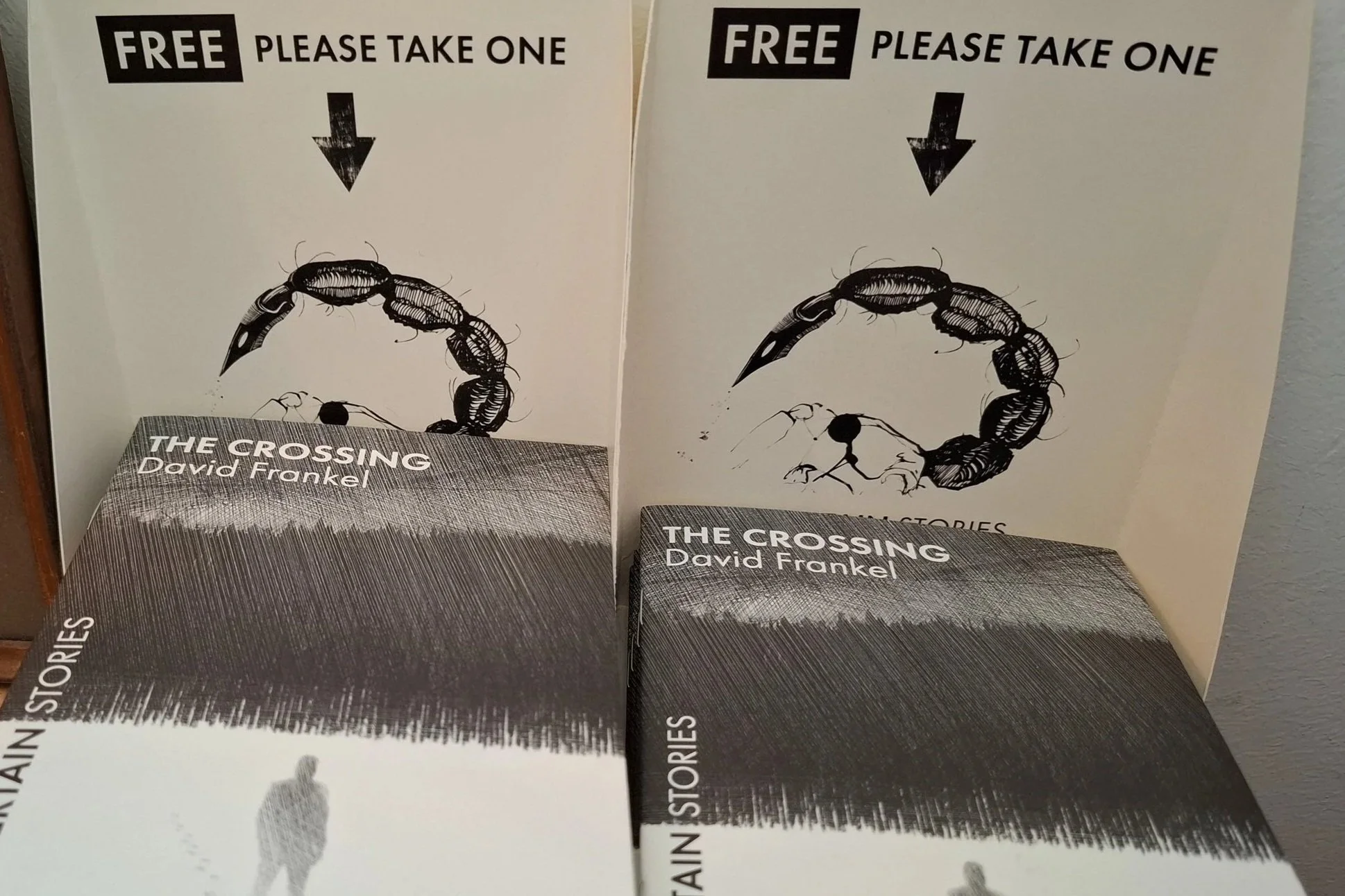 Two book displays featuring the book titled 'The Crossing' by David Frankel. Each display has a sign at the top with the words 'FREE Please Take One' and a black arrow pointing downward. The book cover shows a black and white illustration of a scorpion forming a circular shape.