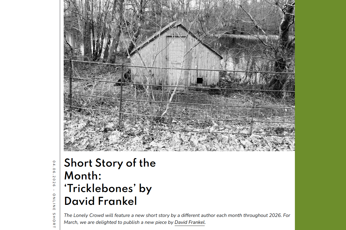 A black and white photo of a wooden shed in a wooded area, surrounded by a metal fence with leaf-covered ground and leafless trees in the background.