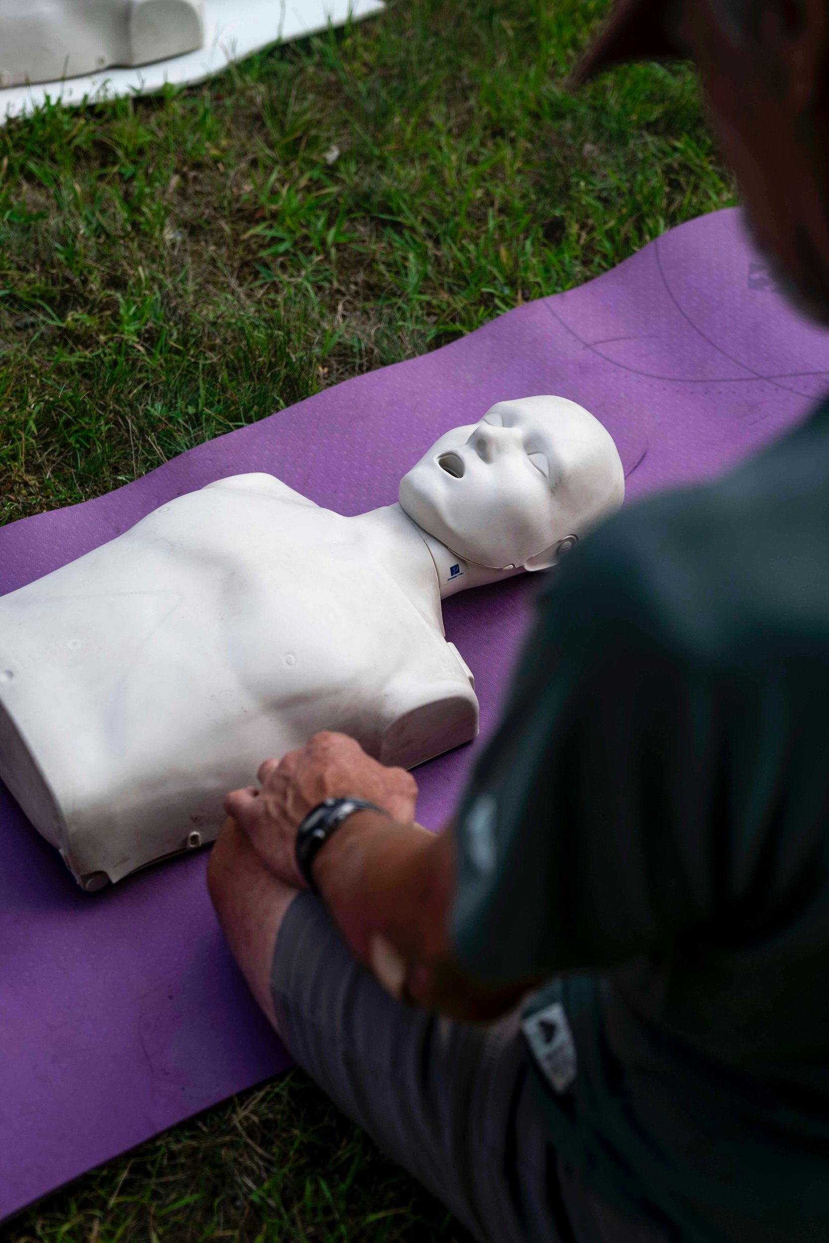 A person demonstrating CPR techniques on a mannequin lying on a purple mat outdoors, with grass surrounding the area.