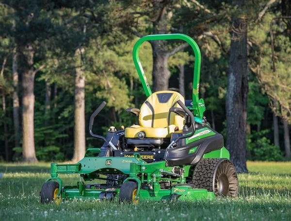 Green and black riding lawn mower with yellow seat on a grassy lawn surrounded by trees.