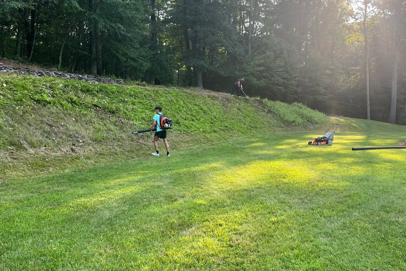 Two people working on a golf course mowing the grass near a wooded area.