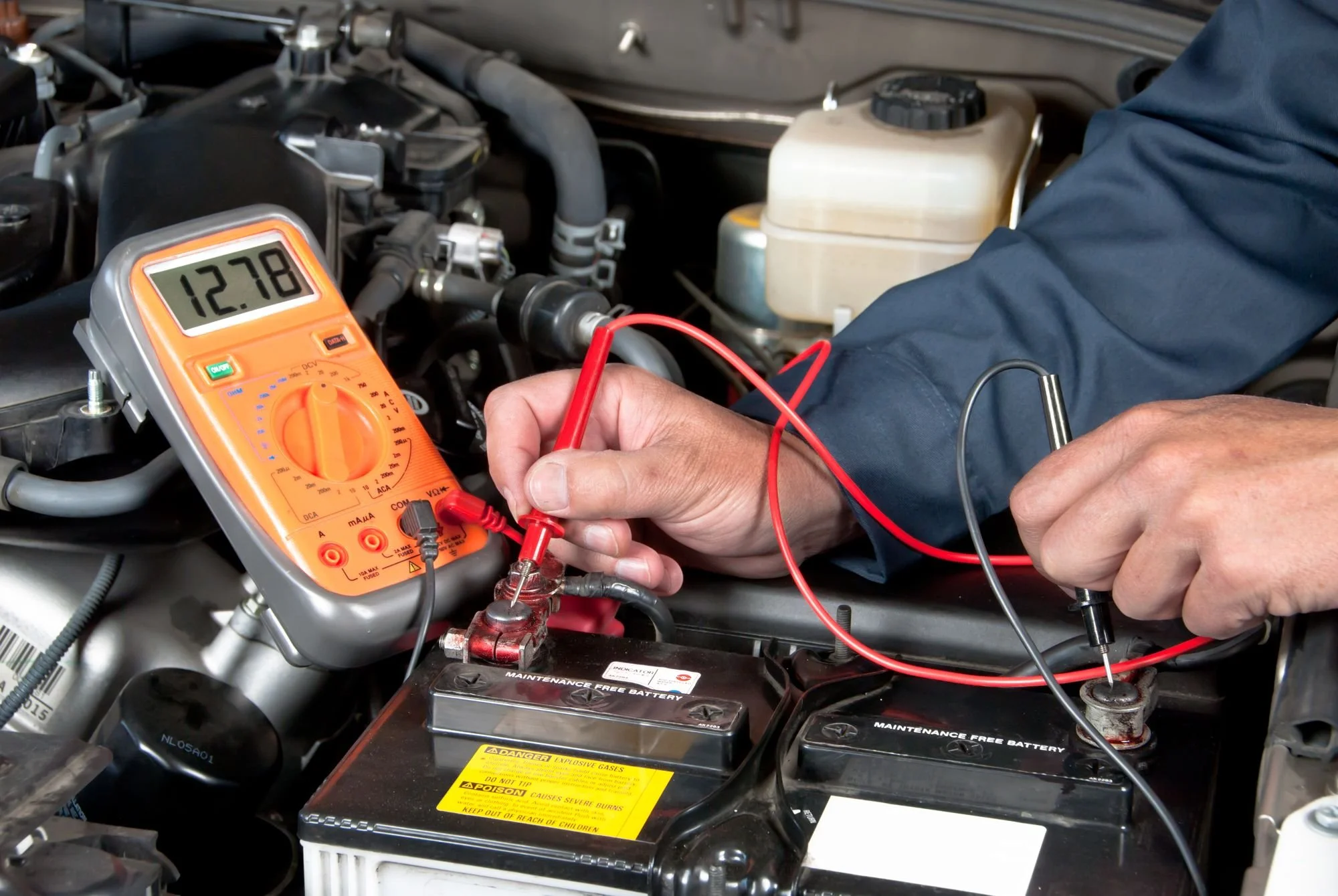 A mechanic using a digital multimeter to test the electrical system of a car engine. The multimeter shows a reading of 12.78 volts. The engine bay contains various components like the battery, coolant reservoir, and hoses.
