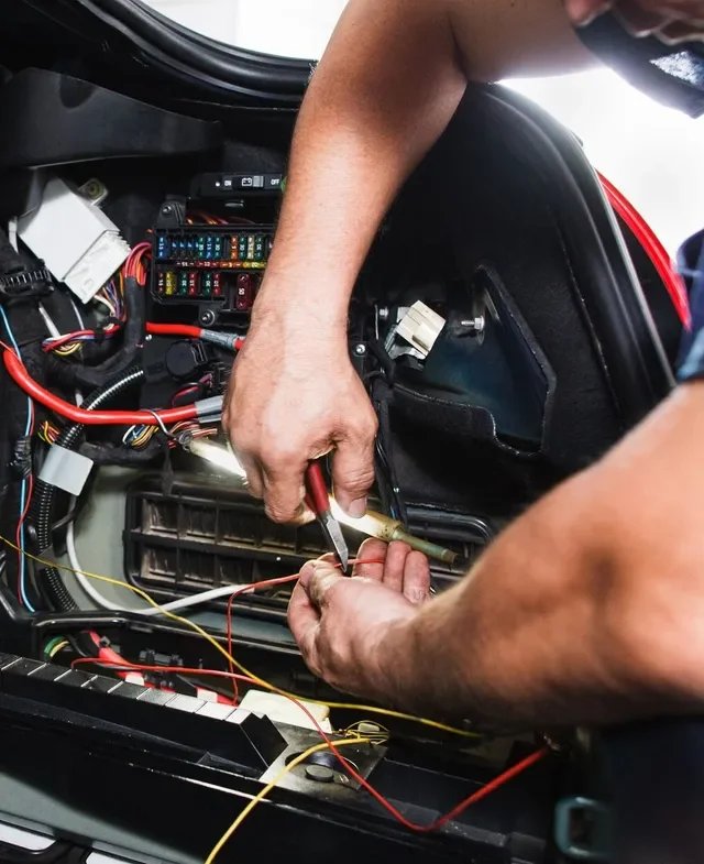 A person working on the electrical wiring inside a vehicle's dashboard or engine compartment, using tools to connect or repair wires.