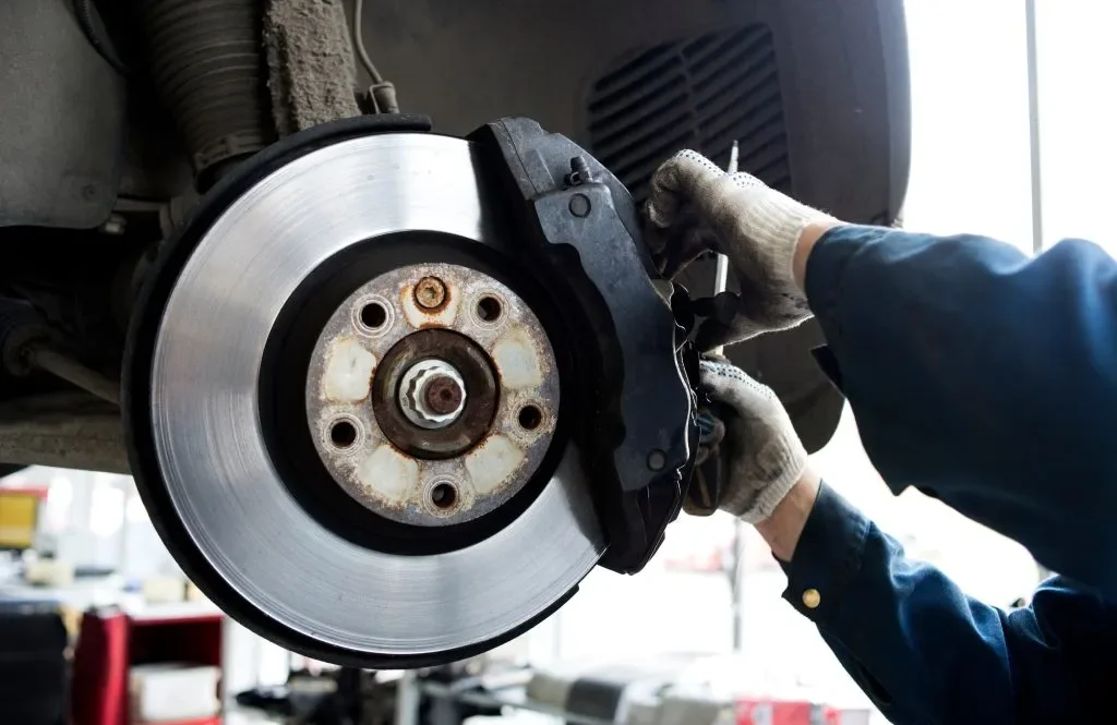 A mechanic wearing gloves and a blue jacket working on a car's brake system at a garage, with the brake rotor and caliper visible.
