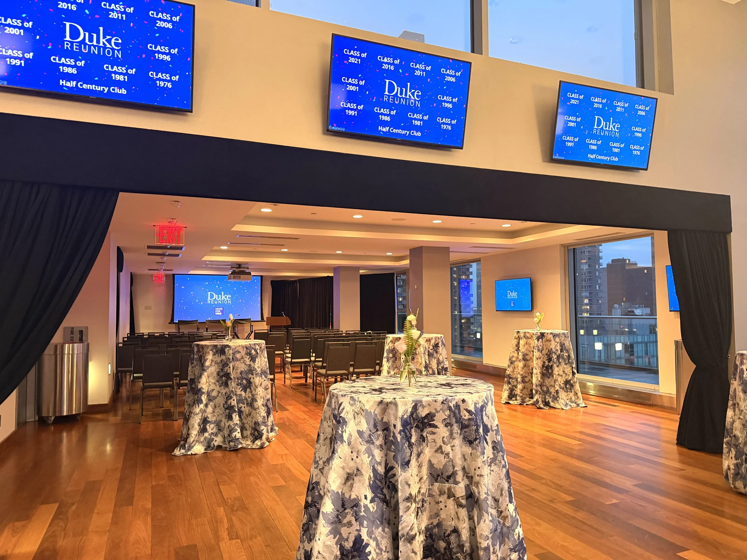 An indoor conference room decorated for a Duke alumni reunion with multiple tuxedo-clad tables draped in floral tablecloths. Mounted screens display a blue graphic for the Duke reunion with class years listed. The room has large windows, wooden floor