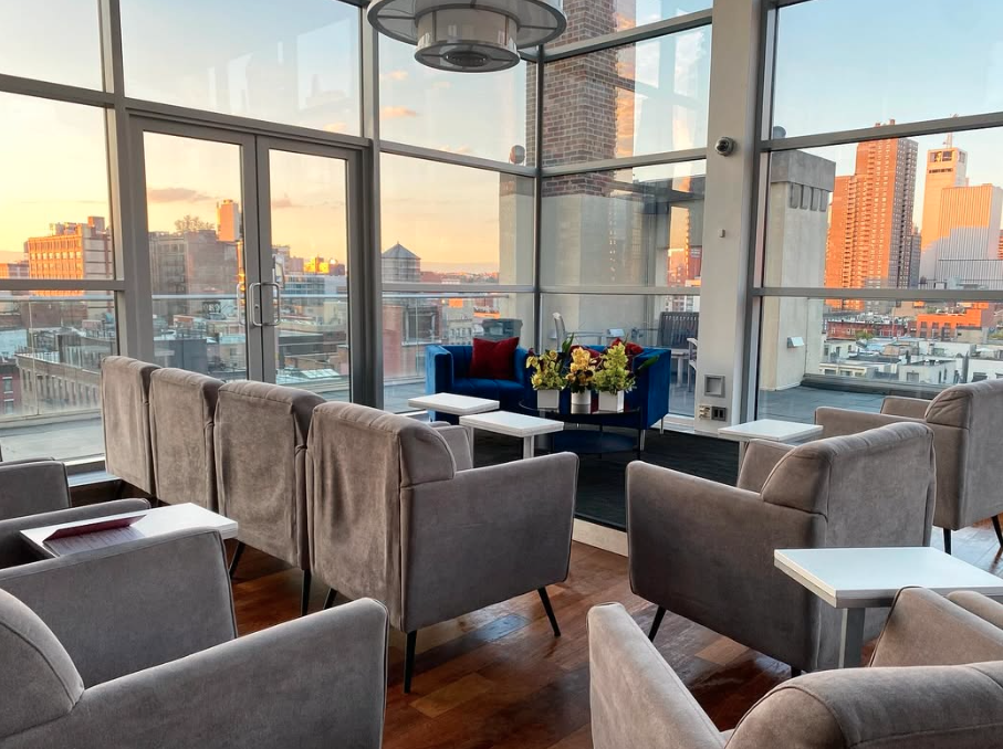 Modern lounge area with large glass windows showing city skyline at sunset, grey upholstered chairs, white side tables, a blue sofa with red cushions, a table with a flower arrangement, and a ceiling light fixture.
