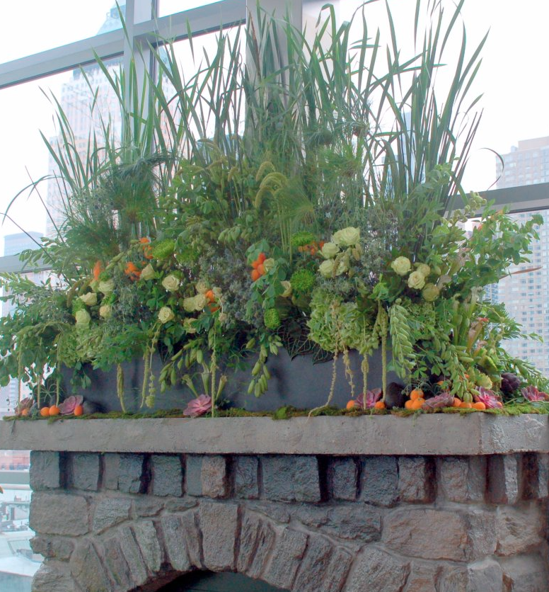 A large flower arrangement on top of a brick fireplace, containing various green plants, white roses, orange flowers, and tall grass or reeds, with large windows and city buildings in the background.
