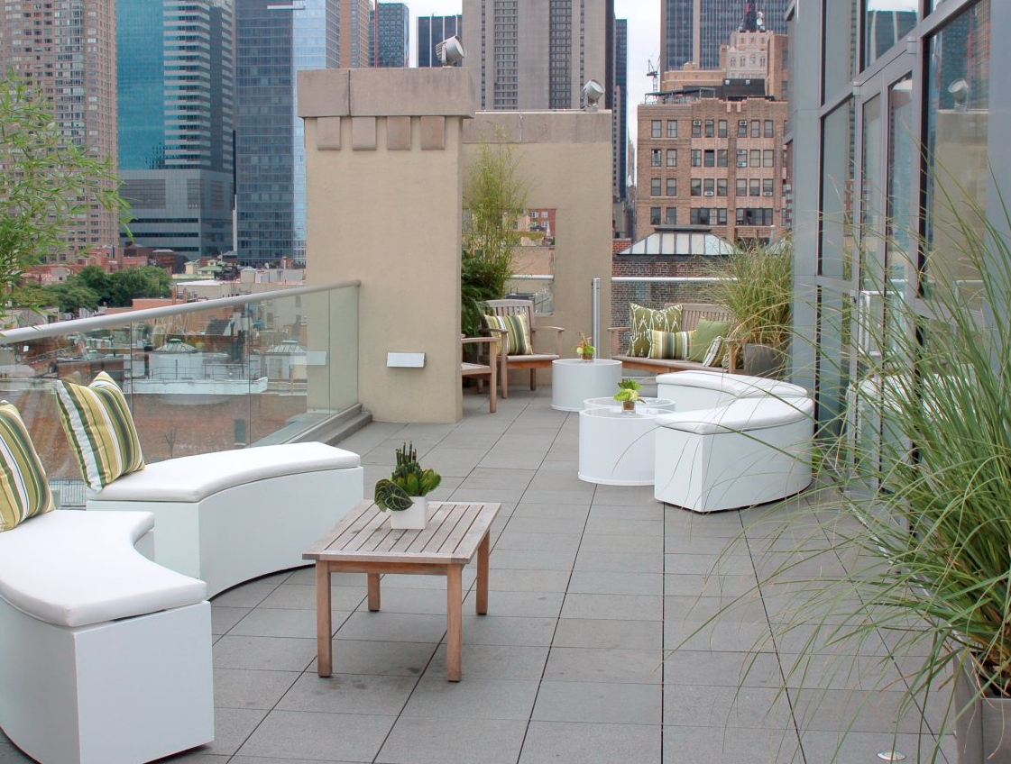 Urban rooftop terrace with white curved sofas, wooden tables, green striped cushions, potted plants, and a city skyline in the background.