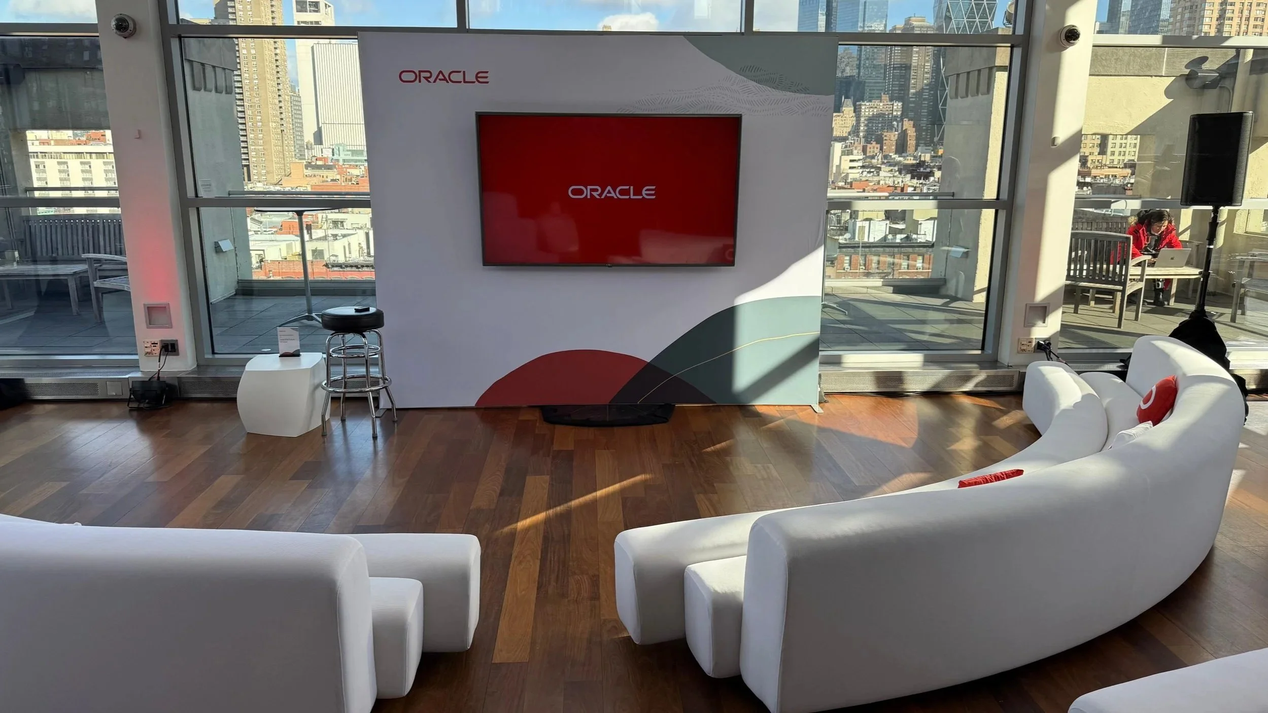 Office event space with a large Oracle logo on the wall, a flat-screen monitor, white couches, a small round table, and floor-to-ceiling windows showing a city skyline. A woman in red is working at a table outside on the balcony.