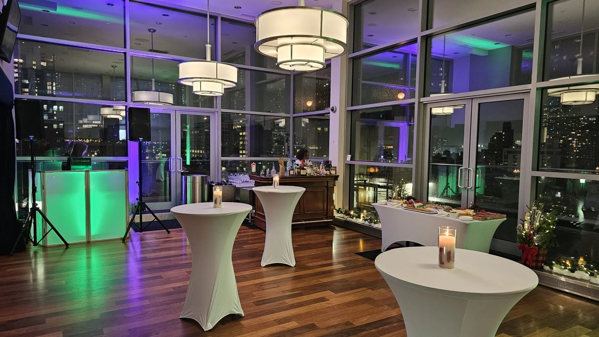 Indoor evening party setup with high cocktail tables, candles, a bar with drinks, and city skyline view through large glass windows illuminated by colorful lights.
