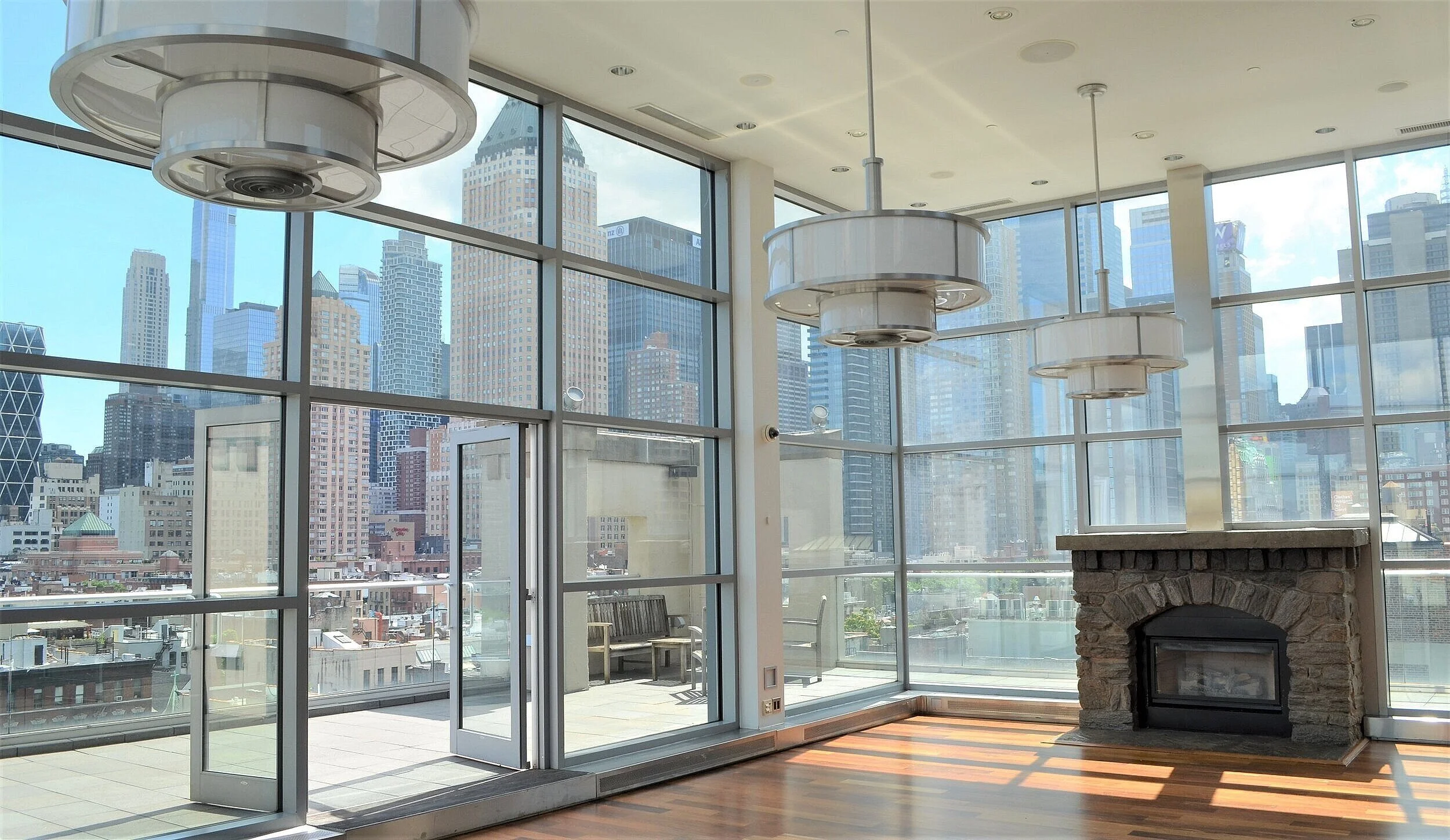Interior of a modern high-rise apartment with large glass windows showcasing a city skyline, a stone fireplace, and ceiling-mounted circular light fixtures.
