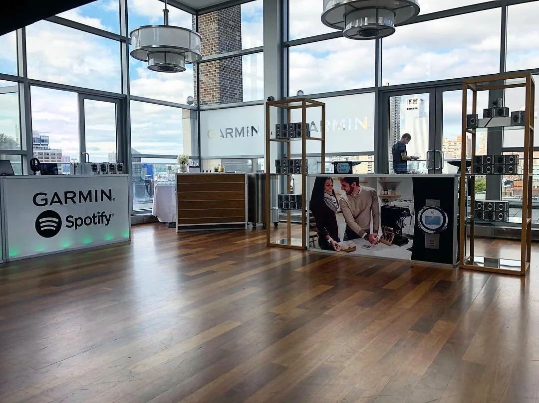 Empty event space with large windows, wooden floors, and displays for Garmin and Spotify, featuring speakers and promotional images.