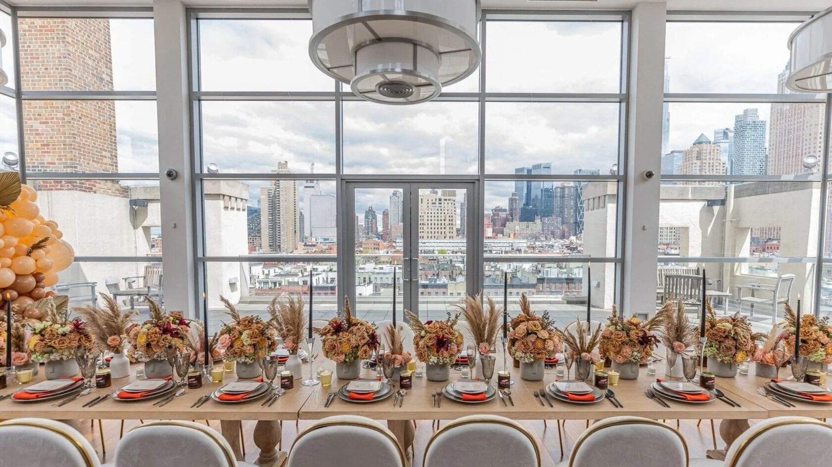 A long table set with floral centerpieces, candles, napkins, and tableware in a bright room with large windows overlooking a city skyline.