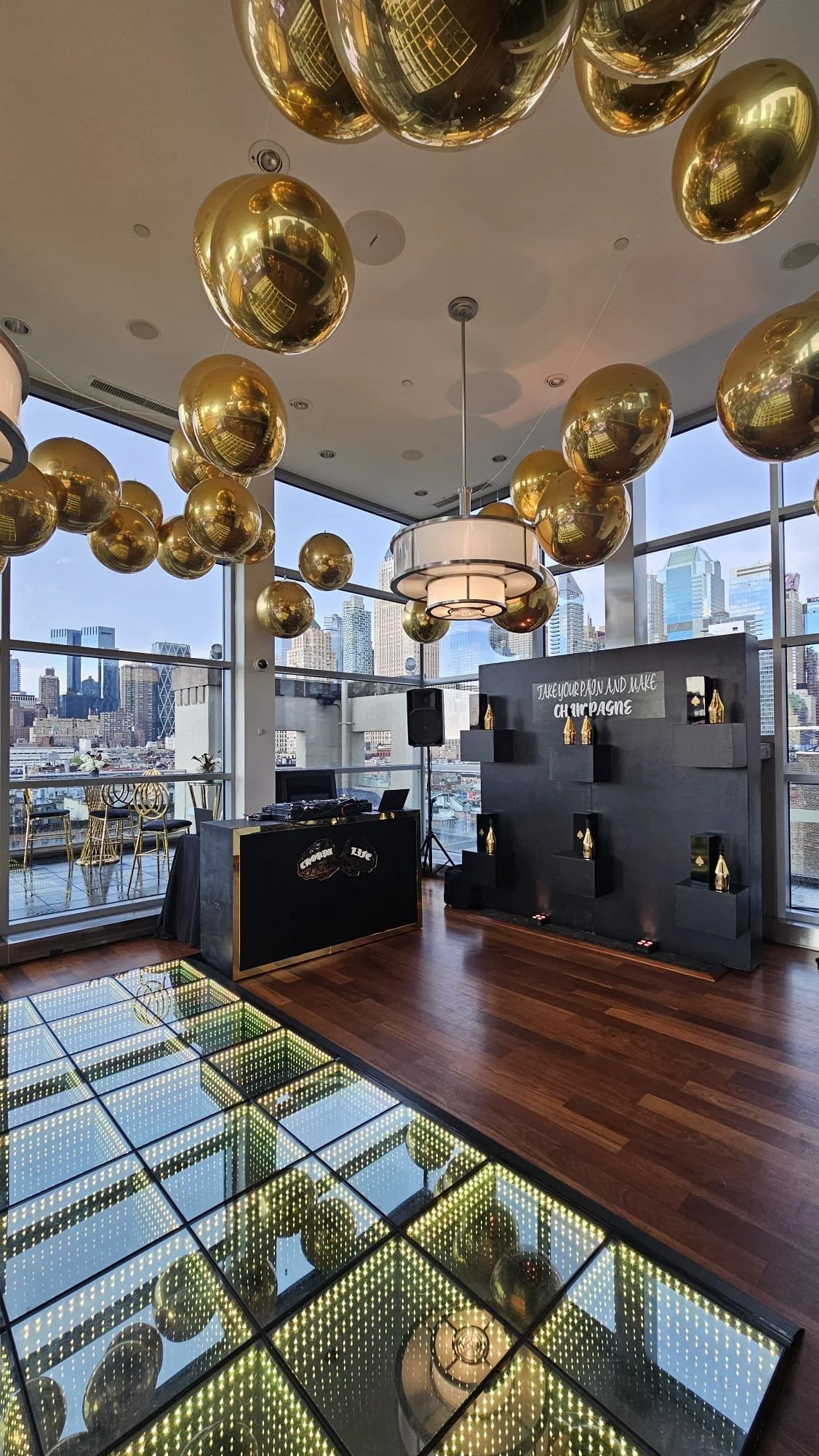 Interior of a modern event space with gold balloons hanging from the ceiling, city skyline view through large windows, a DJ booth, and a wall with champagne bottle decorations.