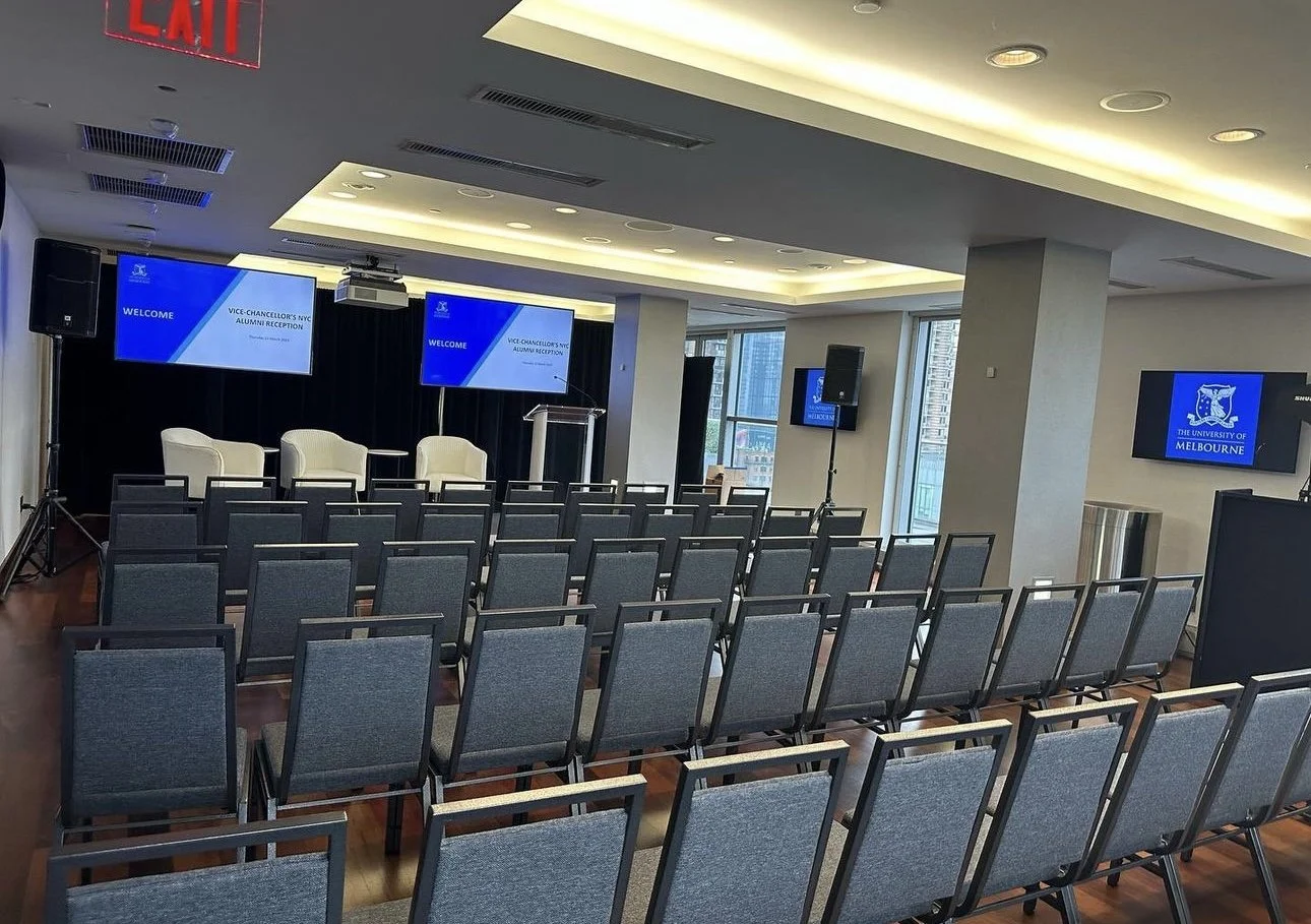 Conference room set up for an event with rows of gray chairs, a stage with three white chairs, and large screens displaying a welcome message for the Vice-Chancellor's NYC Alumni Reception at the University of Melbourne.