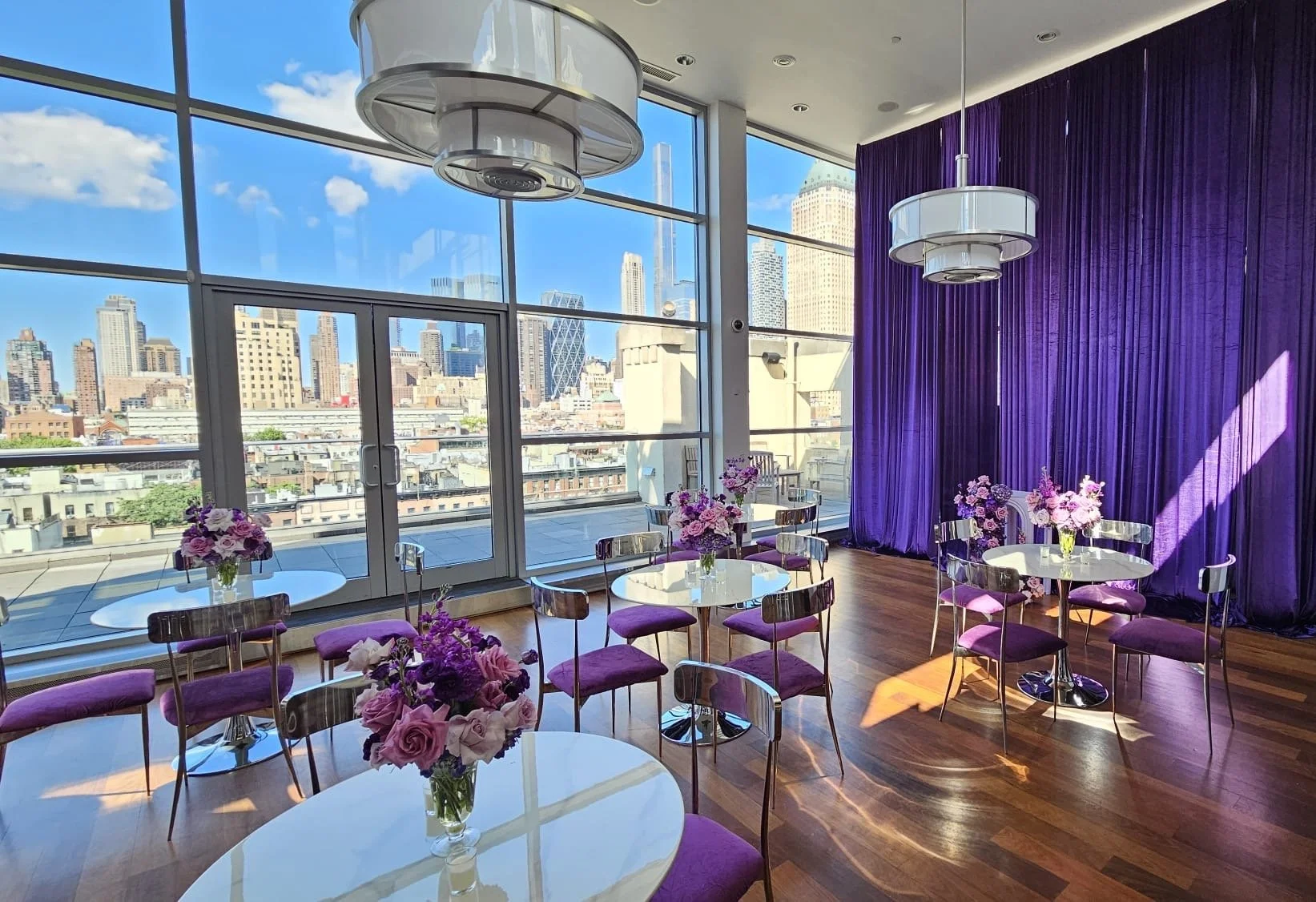 Upscale interior with large windows showing a city skyline, purple curtains, round tables with purple chairs, and vases of pink and purple flowers.