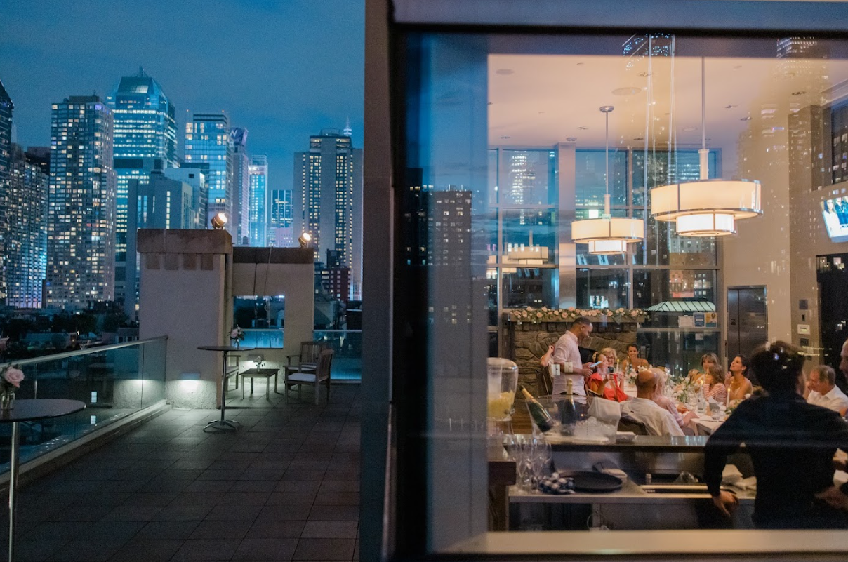 Nighttime cityscape view from a rooftop terrace with tall buildings lit up. Inside, a restaurant with large windows shows people dining and a waiter serving, illuminated by modern hanging lights.
