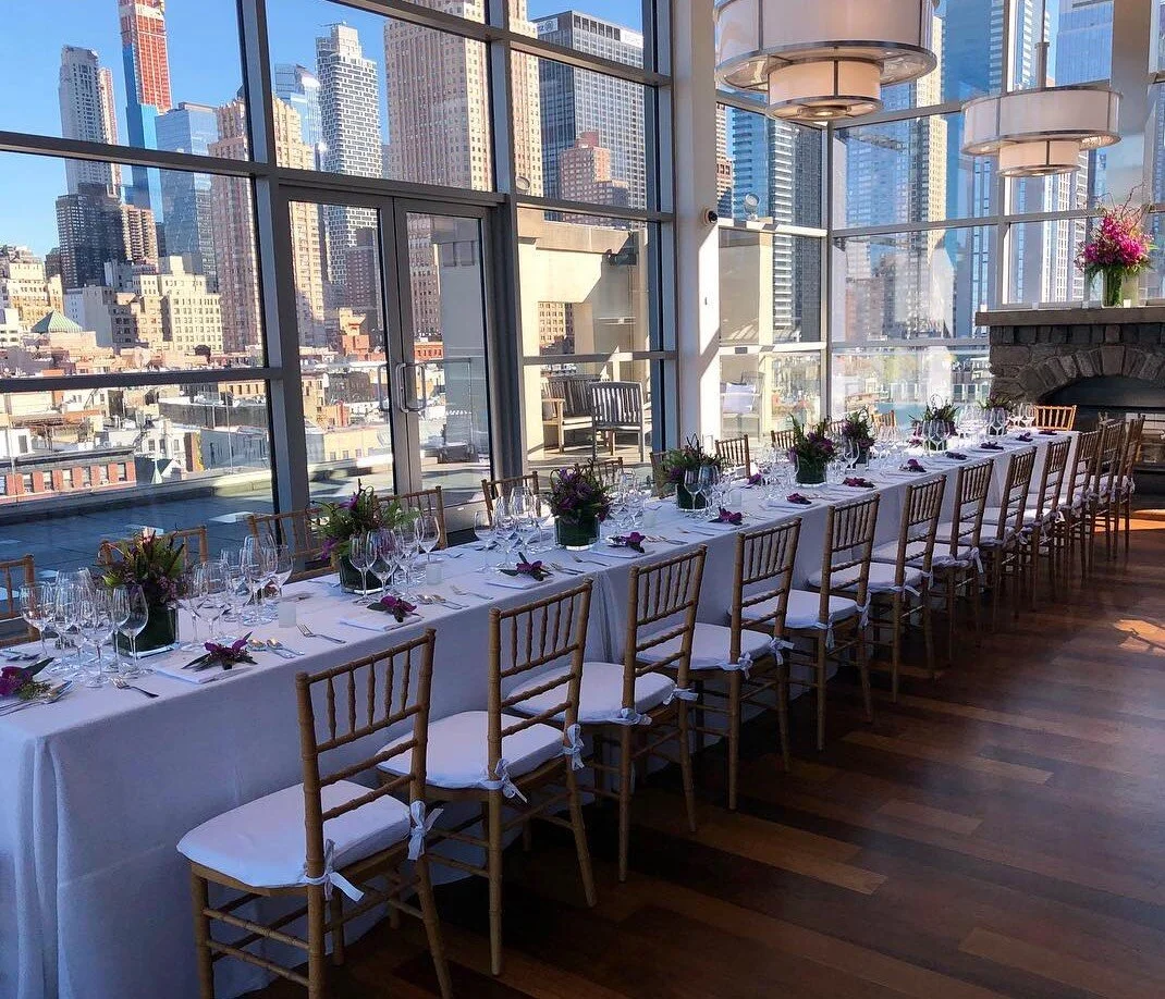 Long banquet table set with plates, glasses, silverware, and purple floral centerpieces inside a bright room with large floor-to-ceiling windows overlooking a city skyline.